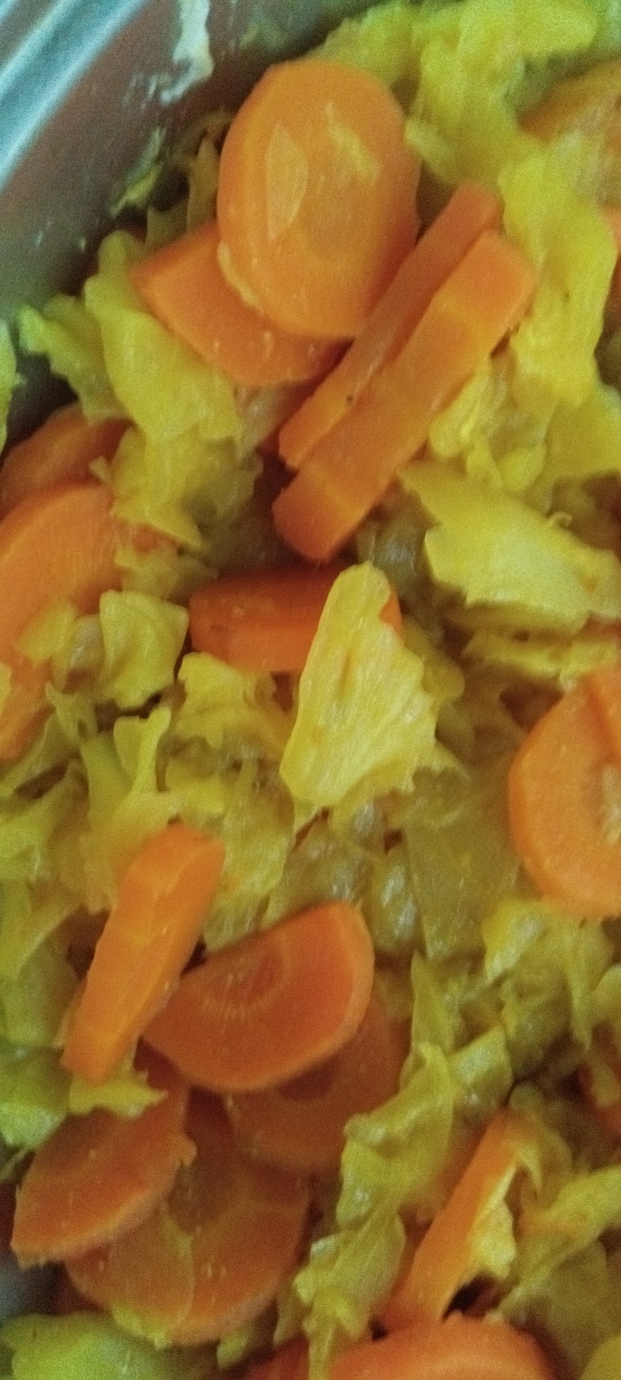 The image shows a close-up, overhead view of chopped carrots and what appears to be cooked cabbage in a metal pot or bowl. The carrots are cut into various shapes, including rounds and diagonal slices, and are bright orange. The cabbage is a pale yellow-green and has a shredded or chopped texture. The lighting is somewhat diffused, and the image has a slight grainy quality. There is no indication of a specific location in Yaroslavskaya, Russia.