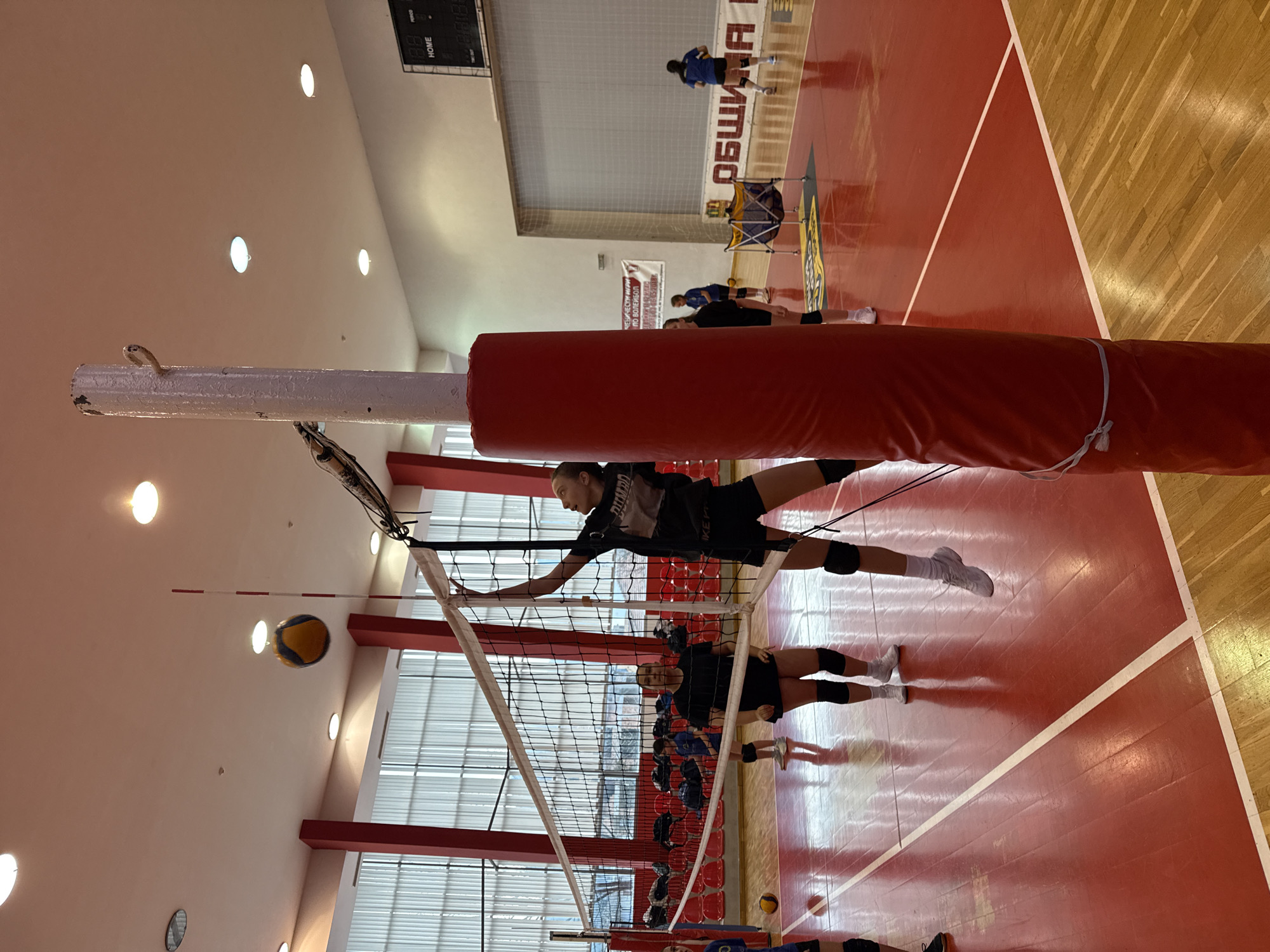 A woman jumps at the volleyball net in a gymnasium with a red and tan floor in Etropole, Bulgaria. She is wearing a black uniform and white socks and has her right arm extended to hit a yellow and blue volleyball. Another woman wearing a similar uniform stands beneath her. The net is white, with a red padded post. There are rows of red seats in the background, and more volleyball players can be seen on the court. Text in Cyrillic letters is visible on the wall. The ceiling has recessed lighting.