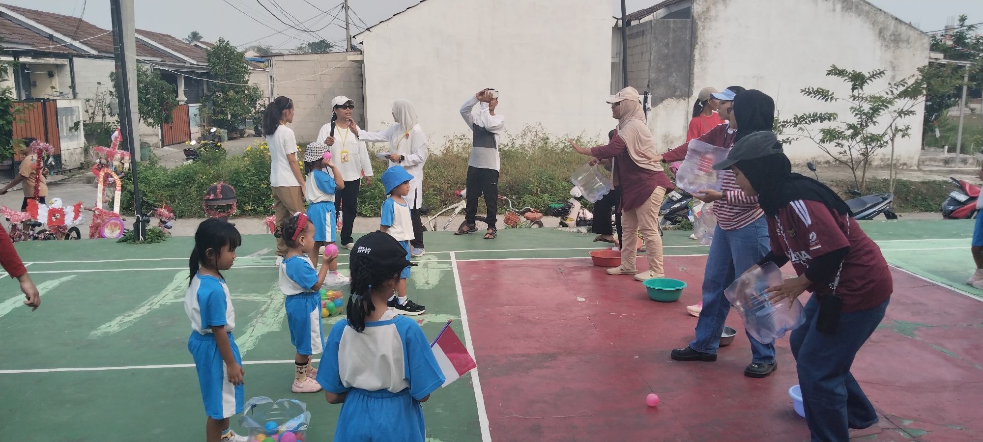 The image depicts an outdoor event taking place on a red and green court, likely a basketball or badminton court, in Taban, Indonesia. Several individuals, including children and adults, are participating in what appears to be a game or activity.
In the foreground, several young children dressed in blue and white uniforms are standing on the court. One child on the far left, facing right, is wearing a blue and white t-shirt and shorts. Next to them, another child, also in blue and white, is looking to the right and holding a pink ball. Further to the right, a child in a blue bucket hat and blue and white outfit is looking forward. Another child, wearing a blue and white uniform and a black cap with "Hello Kitty" on it, is facing away from the camera. The back of another child in a similar uniform, holding a small Indonesian flag, is visible on the far right. A basket filled with colorful balls is placed near this child.
In the middle ground, several adults are interacting with the children. One adult, wearing a white shirt and black pants, is holding a microphone and speaking to the children. Another adult in a white shirt and white cap is also present. To the right, an adult in a tan hijab and a maroon shirt, holding a large plastic bottle, is looking towards the children. Another adult in a red shirt and a cap is also present. Several adults in the background are also engaged in the activity, some holding plastic bottles.
The environment shows houses with tiled roofs in the background, suggesting a residential area. There are also trees and vegetation surrounding the court. Various objects, including bicycles, bags, and buckets, are scattered around the edges of the court. The sky is overcast, indicating a cloudy day. The overall scene suggests a community event or a school activity taking place outdoors.
