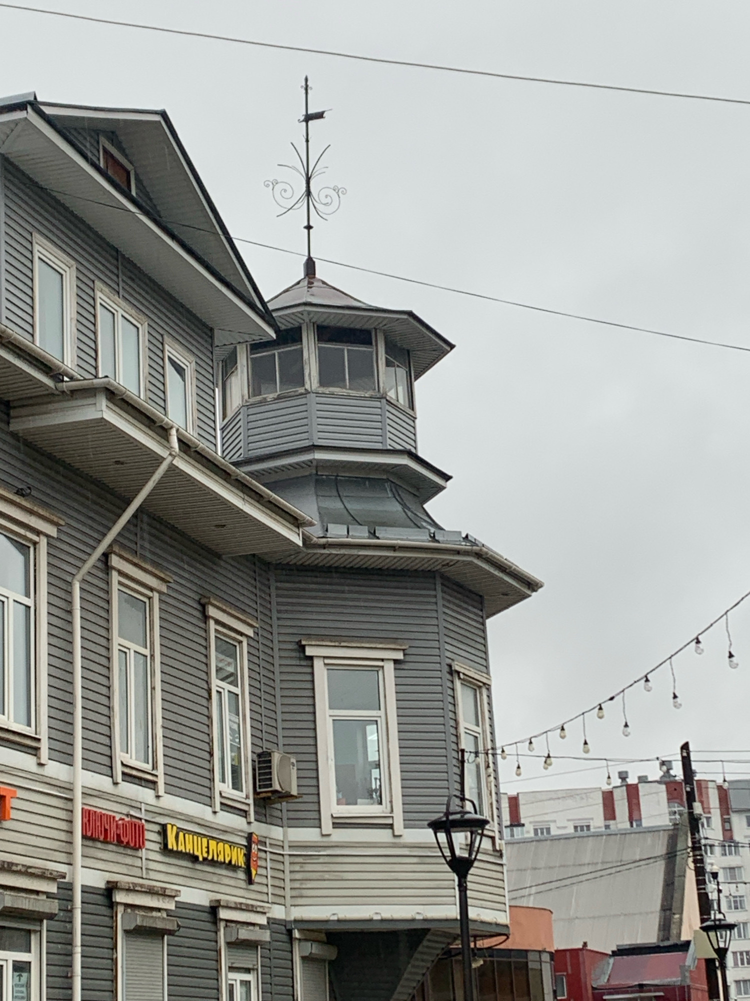 The image shows a close-up of a gray, wooden building with multiple windows and a decorative weather vane on the roof. The building appears to be an older structure, possibly with commercial establishments on the ground floor, indicated by signs that read "КЛЮЧИ-ФОТО" (Keys-Photo) and "КАНЦЕЛЯРИК" (Stationery). The building has a distinctive octagonal turret with windows. In the background, other buildings and a cloudy sky are visible, along with strings of lights that are likely for decoration. The setting is urban, and the overall impression is that of a street scene in a city. Based on architectural style and the Russian text on the signs, it is likely located in Russia, possibly Arkhangelsk. There are no people visible in the image, and the focus is entirely on the building and its immediate surroundings.