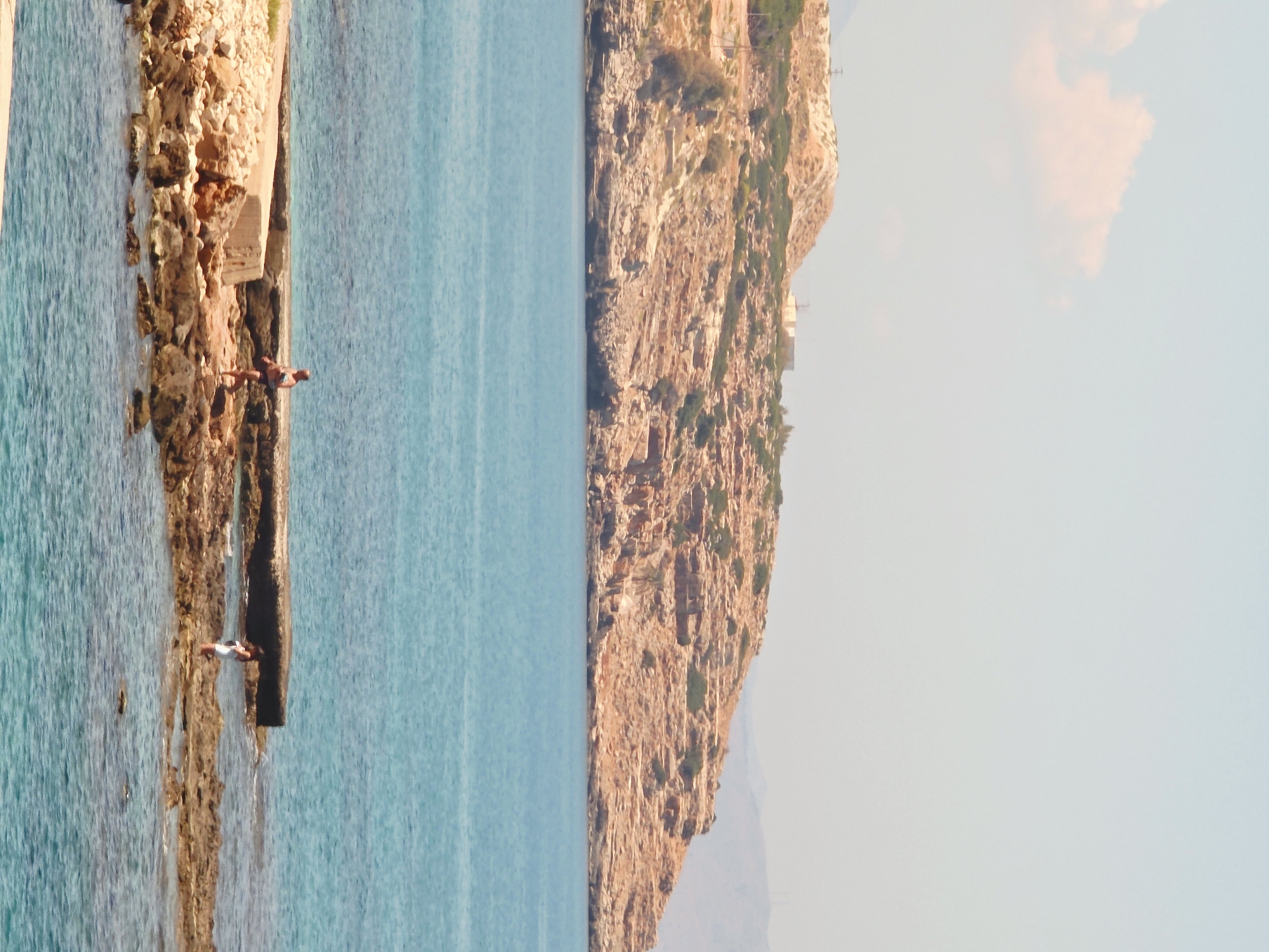 The image depicts a rocky coastline with clear blue water. In the foreground, two individuals are visible on the rocky shore. One person, appearing to be a male, is standing and looking to the left. Another person, appearing to be female, is standing near the water's edge, facing right. In the background, a rocky, arid hill rises from the sea. The sky is a pale blue with a few scattered clouds. This scene is likely located in or near Rethymno, Greece, given the characteristic Mediterranean landscape.