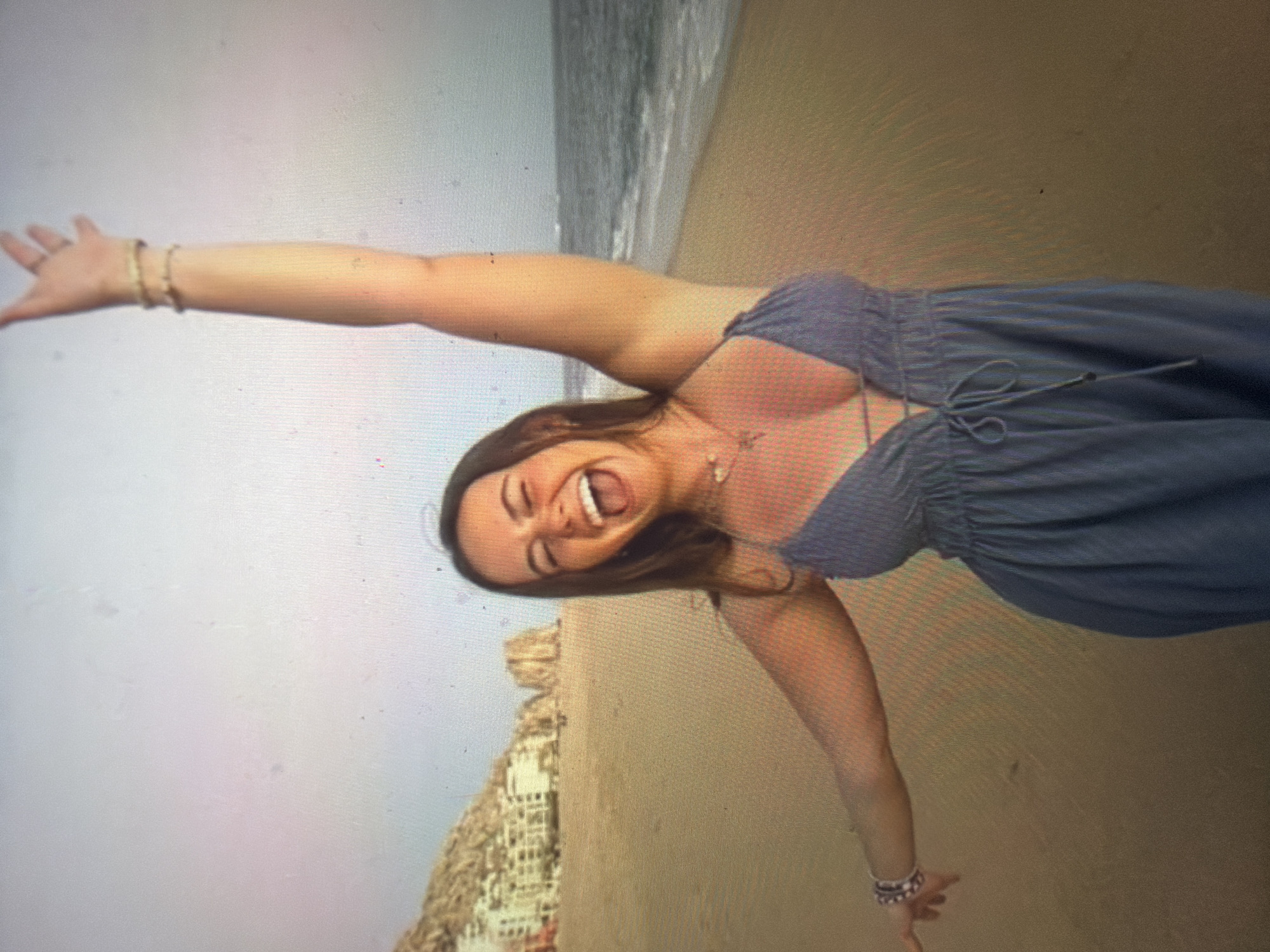 A young woman with long brown hair, wearing a blue sleeveless garment with a V-neckline and tie detail, stands on a sandy beach. Her left arm is raised high with an open hand, displaying gold-toned bracelets, while her right arm is extended to her side, also adorned with a bracelet. She is captured mid-laugh, with her mouth wide open and eyes squinted in joy. A small, heart-shaped pendant hangs from a necklace on her chest. The background features a vast body of water with visible waves on the right side. To the left, a series of large, light-colored rock formations and cliffs rise from the ocean, upon which several multi-story, light-colored buildings are constructed. The sky above is bright and appears slightly hazy. This coastal scene is located in the city of Shumen, Bulgaria.