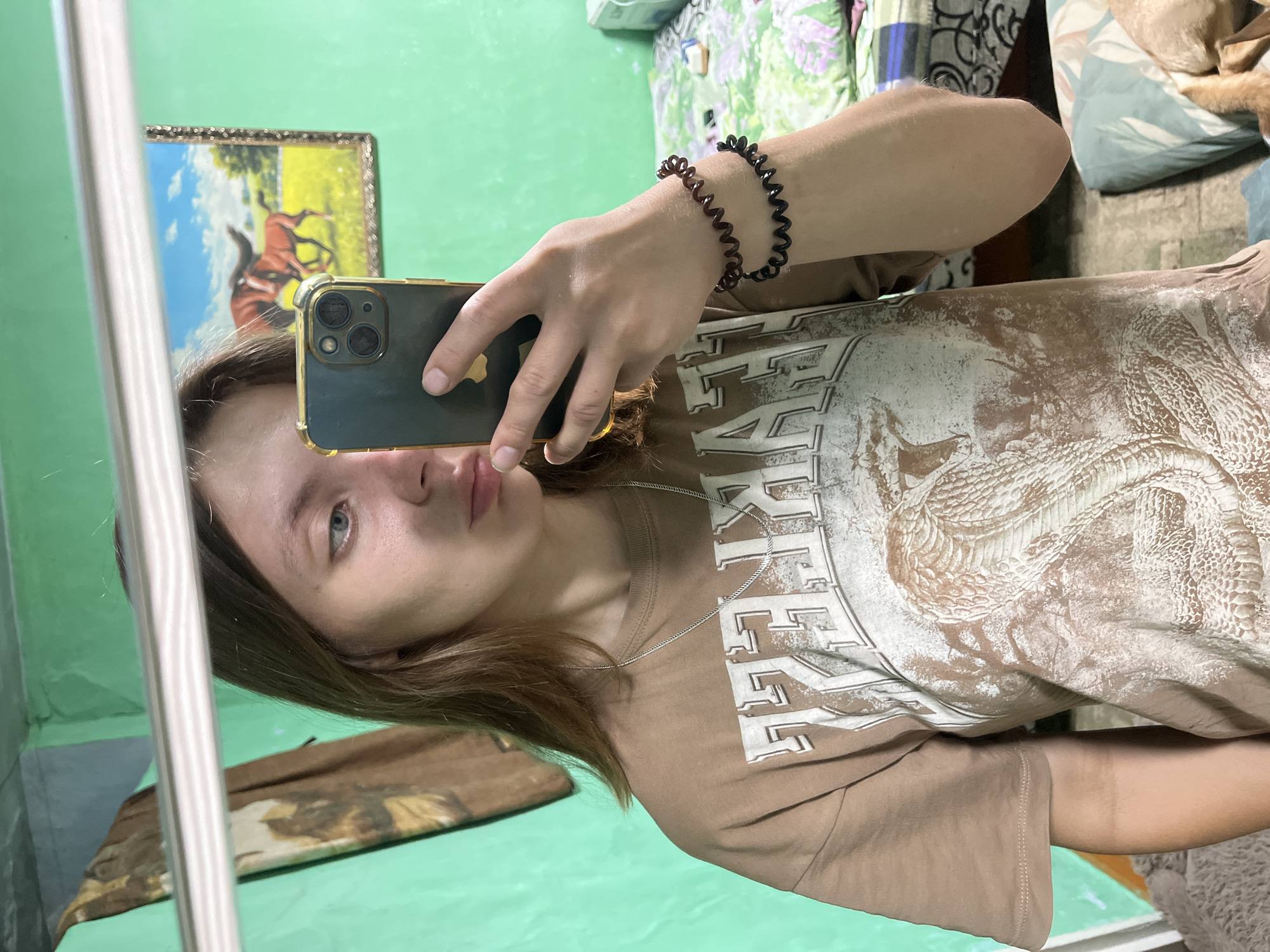 A young woman with light brown hair and blue eyes is taking a selfie in a room with green walls. She is wearing a brown t-shirt with a graphic print of a snake and what appears to be text. She has a silver chain necklace and two spiral-shaped bracelets on her left wrist. In the background, a painting of two horses is visible on the wall, as well as a bed with a patterned comforter. The scene is consistent with an indoor residential setting, possibly a bedroom, in Nizhny Tagil, Russia.