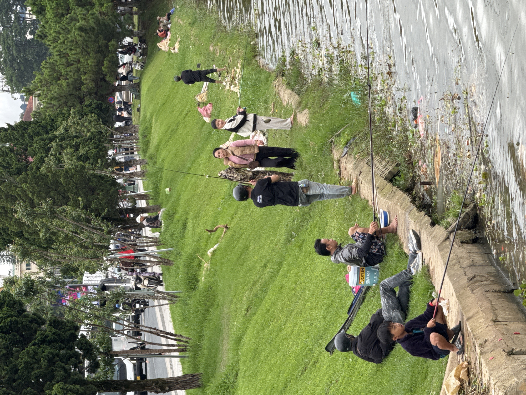 The scene depicts a group of people fishing along the edge of Xuan Huong Lake in Da Lat, Vietnam. A grassy bank separates a street with parked motorbikes from the lake. Several individuals are positioned along the lake's edge, some seated and others lying down, each holding a fishing rod. The lake's surface is visible with reflections and some debris. The setting includes trees on one side, and the street with vehicles on the opposite side, indicating an urban environment adjacent to a natural body of water.