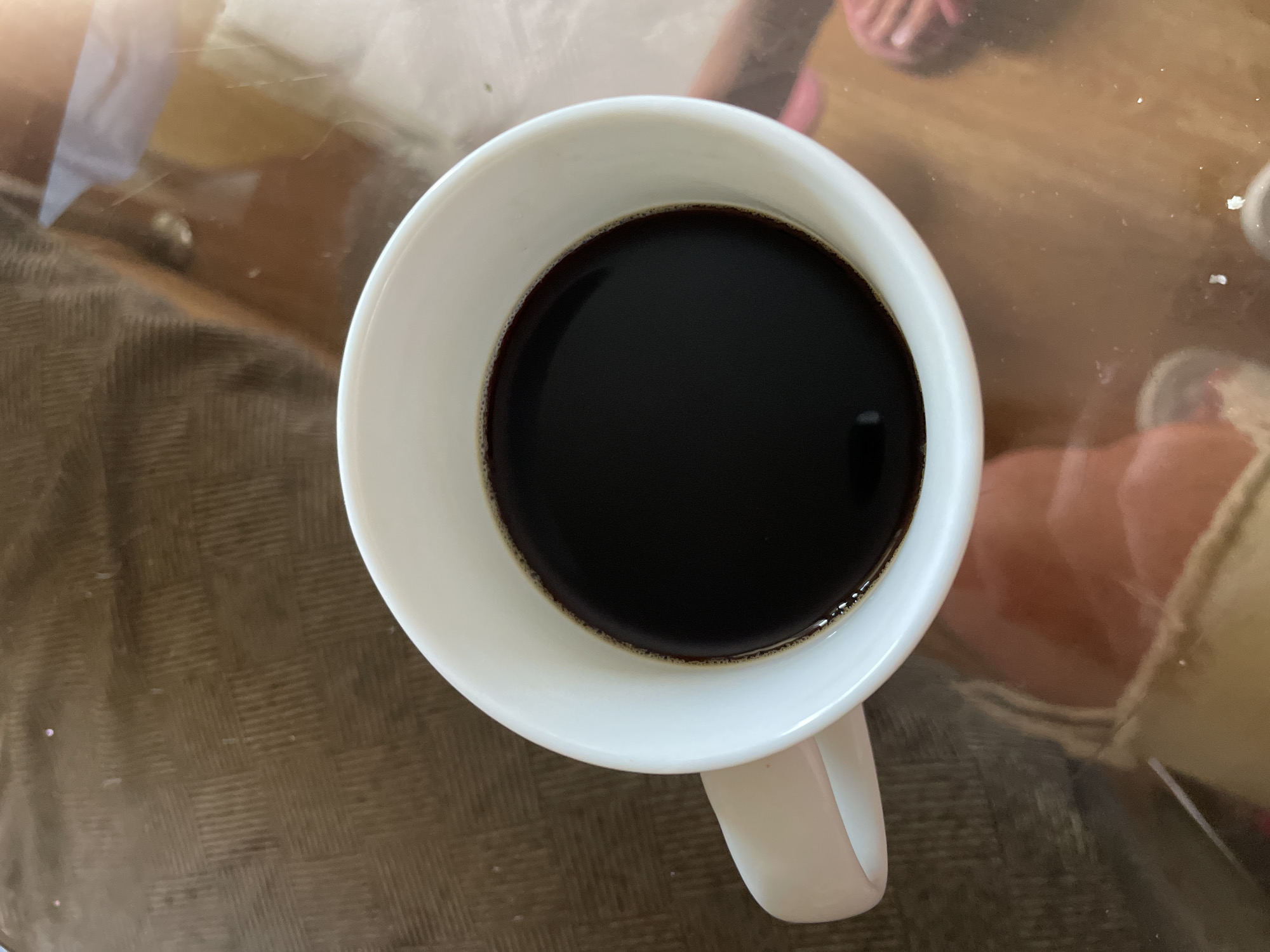 The media file shows a top-down view of a white mug filled with dark coffee. The mug is placed on a textured surface, possibly a table with a placemat or tablecloth. The coffee has a slight sheen and appears to be freshly brewed. The handle of the mug is visible on the right side.
In the background, reflections on a glass surface suggest an indoor setting. There are blurred hints of people, with what appears to be feet wearing pink sandals visible in the upper right corner. The lighting suggests it could be daytime, possibly morning, given the presence of coffee. The overall scene is casual and domestic. There is no visible text in the image. The location context is noted as Kyiv, Ukraine, but there are no specific visual cues within the image to confirm this.