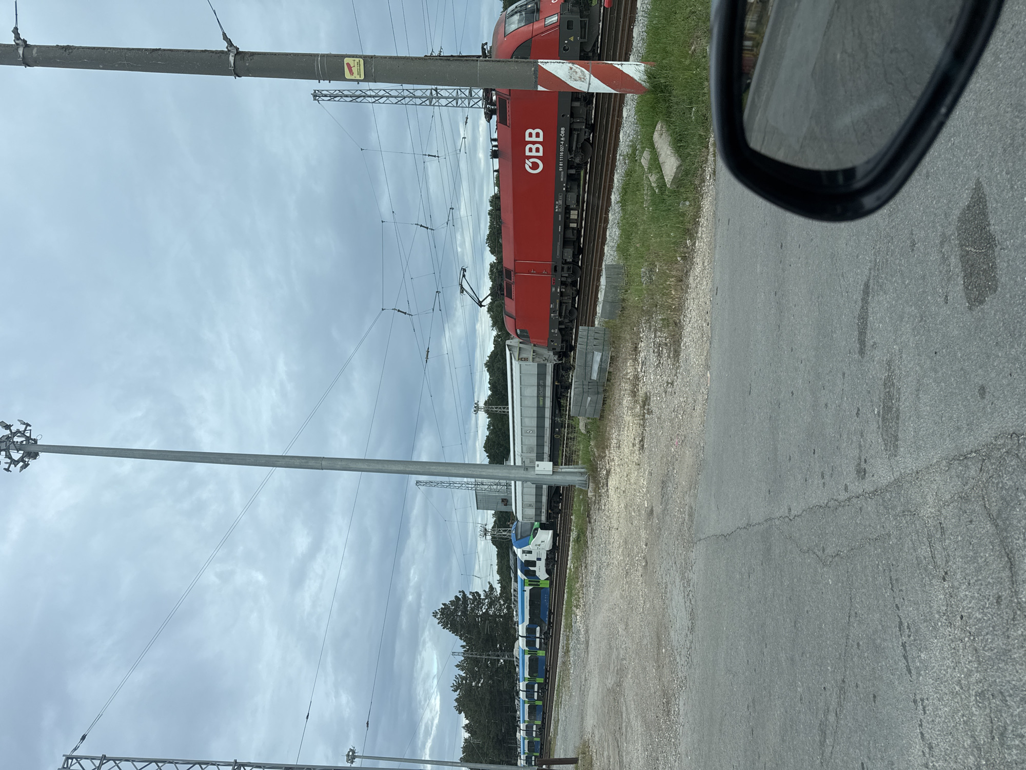 The image shows a train crossing in Senovo, Bulgaria. An ÖBB (Austrian Federal Railways) red electric locomotive is visible on the tracks, coupled with other rail cars. The crossing is adjacent to an asphalt road, with a vehicle side mirror visible in the frame suggesting the photo was taken from a car. Overhead power lines and support structures are present. The sky is overcast. A multi-unit passenger train sits on a different track from the red electric engine.