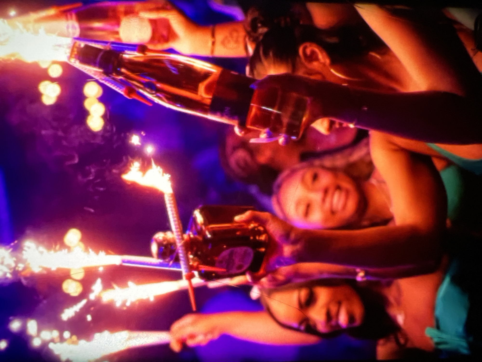 In a dimly lit, vibrant nightclub setting, possibly in Lagos, Nigeria, a group of individuals are celebrating. The foreground is dominated by several people, their faces partially obscured by the low light and the energetic atmosphere. In the center, a person holds up a dark-colored bottle, possibly alcohol, with what appear to be sparklers attached to it. These sparklers are emitting bright, fiery trails of light, adding to the festive ambiance. Behind this central figure, other individuals are visible, some with their arms raised. The overall impression is one of a lively celebration, with a focus on the bottle and the pyrotechnic display. The colors in the image are saturated, with strong contrasts between the bright lights of the sparklers and the deep shadows of the club. The environment appears to be crowded, typical of a nightlife venue.