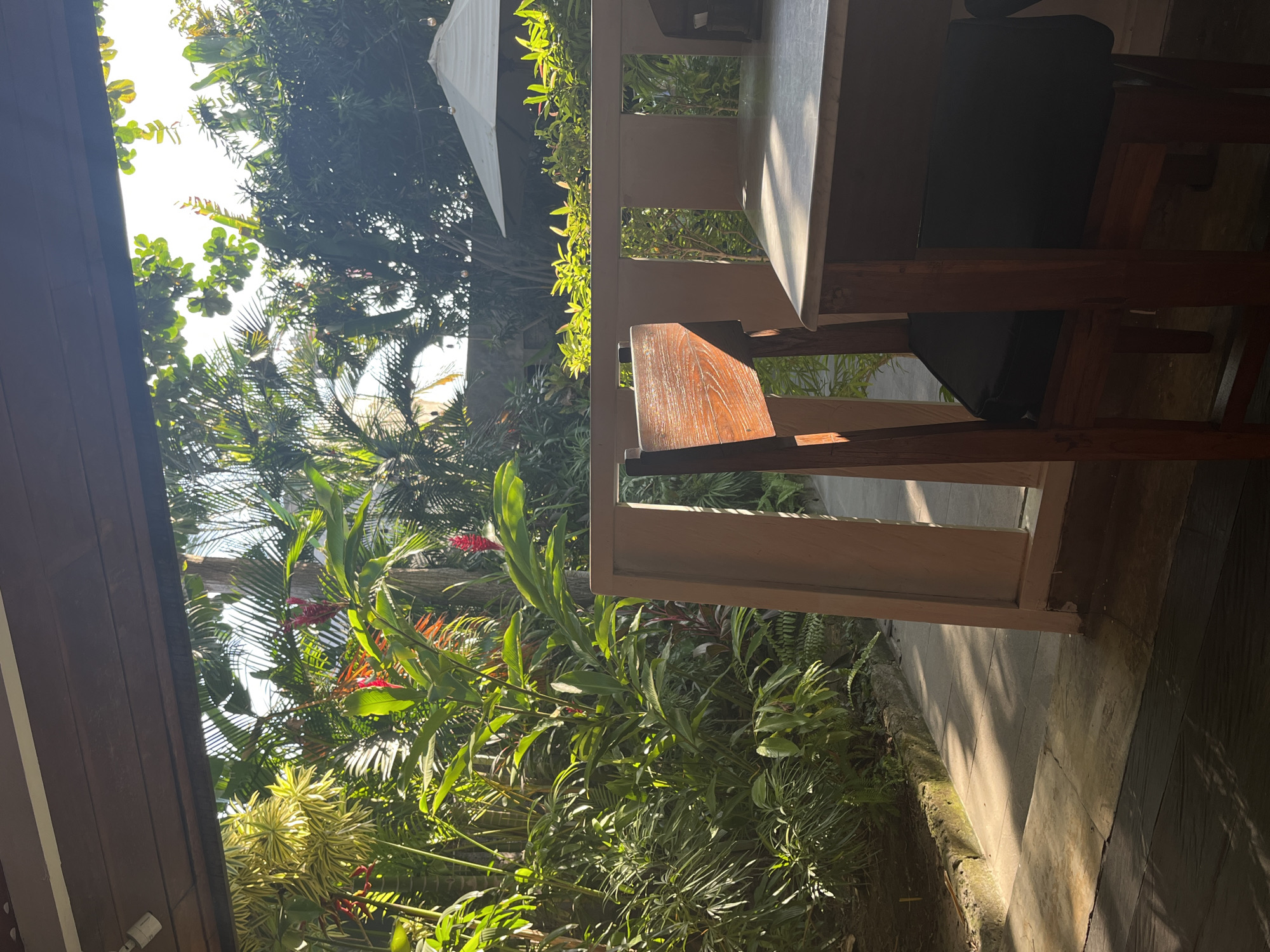 A wooden chair with a black cushion sits at a marble-topped table in an outdoor setting. Lush green foliage with red flowers, palm trees, and sunlight filtering through leaves are visible in the background. A white umbrella is also present in the distance. The scene suggests a tranquil, tropical environment, possibly a cafe or restaurant patio in North Kuta, Indonesia.