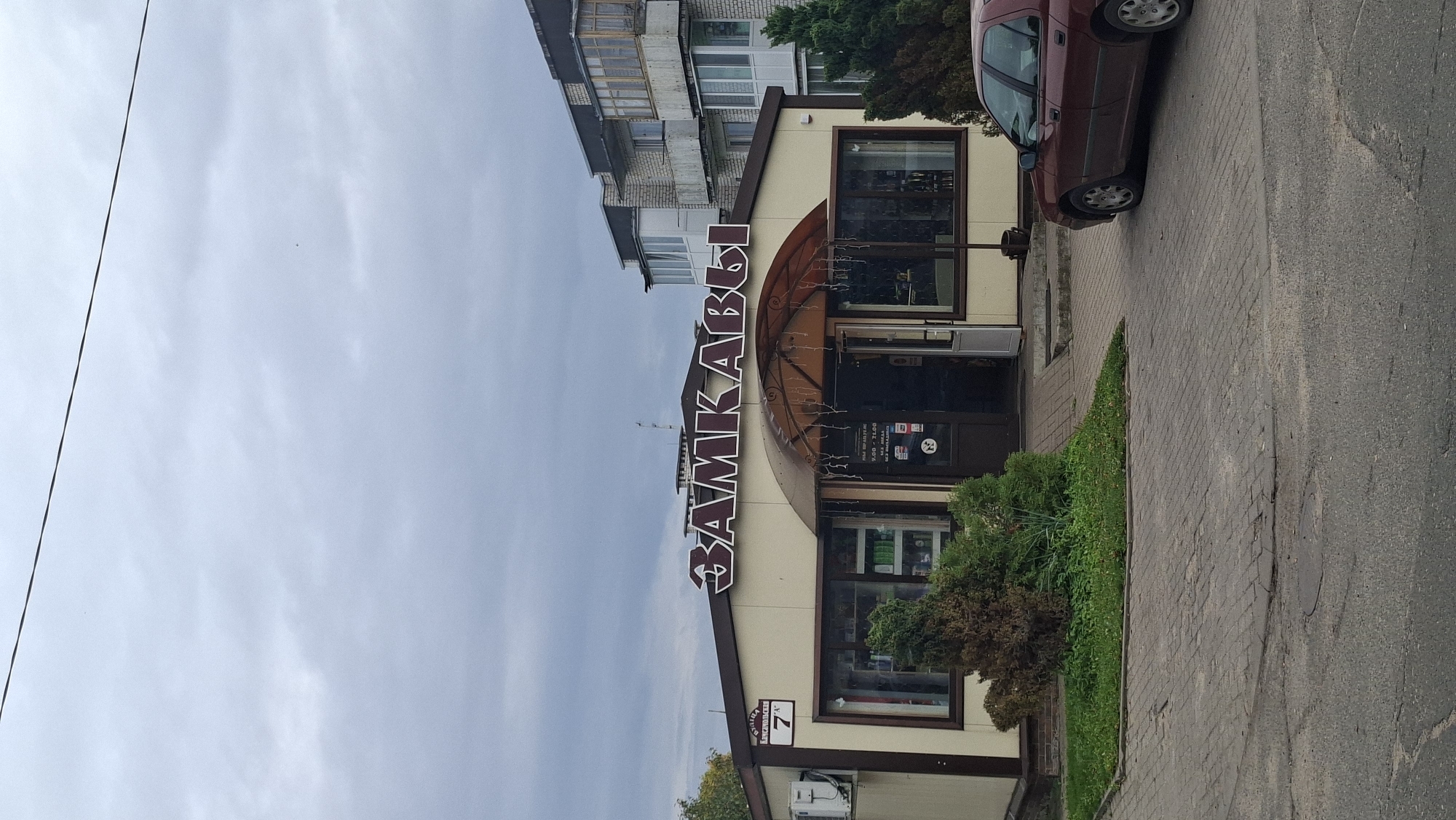 The image shows a storefront with a large sign above the entrance that reads "ЗАМКАВЫ" in large, reddish-brown letters. The building is light-colored with dark brown trim around the windows and entrance. To the left of the entrance, a sign with the number "7" is visible, along with some text in Cyrillic script, indicating it's likely a street address in Myadzyel, Belarus. The storefront has large windows displaying products, and a small patch of green landscaping with a few small bushes is in front of it. An awning with decorative lights covers the entrance. A dark red sedan is parked to the right of the building. The sky is overcast with clouds.