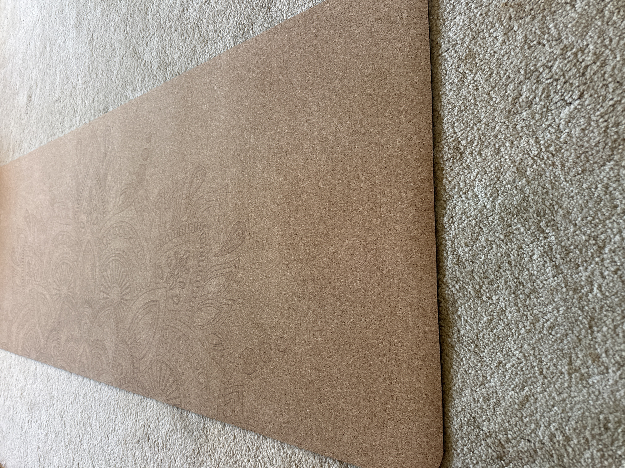 The image shows a close-up, high-angle view of a tan yoga mat with a patterned texture, lying on a beige carpet. The yoga mat is positioned diagonally across the frame, with its surface angled towards the upper right. The pattern on the mat appears to be a paisley or floral design etched into the material. The carpet has a shaggy texture. The scene is lit by natural light, casting subtle shadows. No people or specific locations in the city of Thora, Australia, are visible in the image.