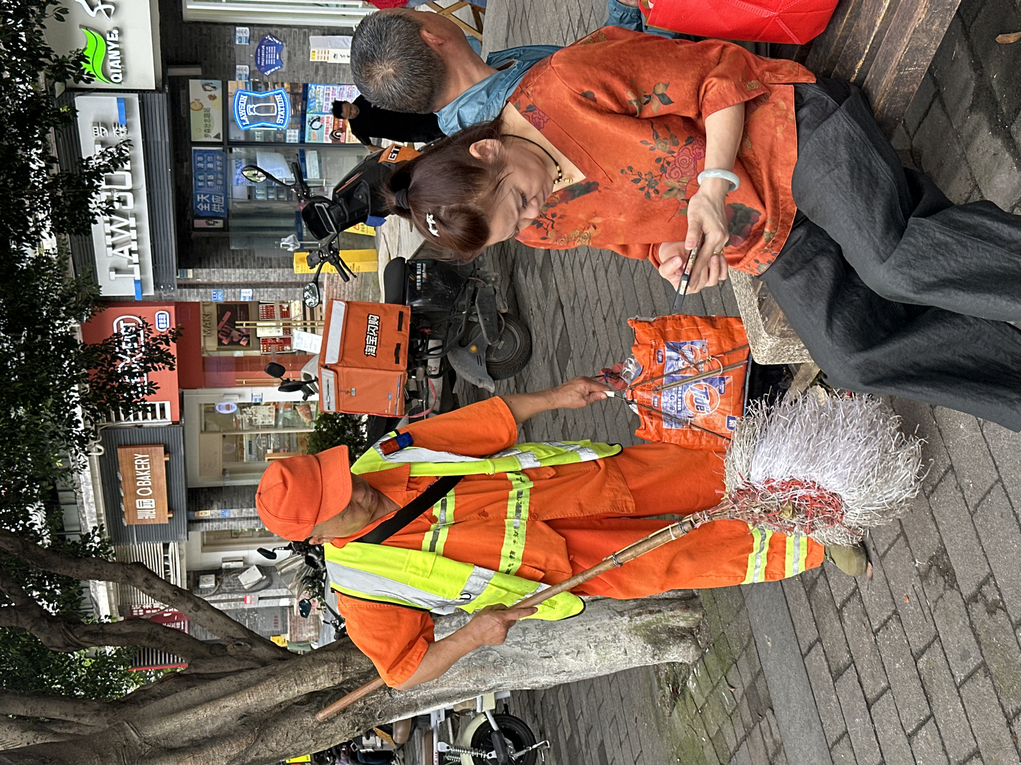 The image captures a scene in Shapingba District, China, depicting an interaction between a sanitation worker and a woman. The woman, wearing an orange floral top and black pants, sits on a bench while using her phone. To her left is an older man with gray hair and a blue jacket. The sanitation worker, dressed in an orange uniform with reflective yellow stripes and a matching cap, stands next to the bench with a broom. He appears to be holding out a small item toward the woman, possibly a piece of trash. Behind them, a motorbike with a delivery box is parked. In the background, there are several storefronts with Chinese signs, including "LAWSON STATION", "QIANYE", and "Q BAKERY". The street is paved, and a tree with exposed roots is visible on the left.