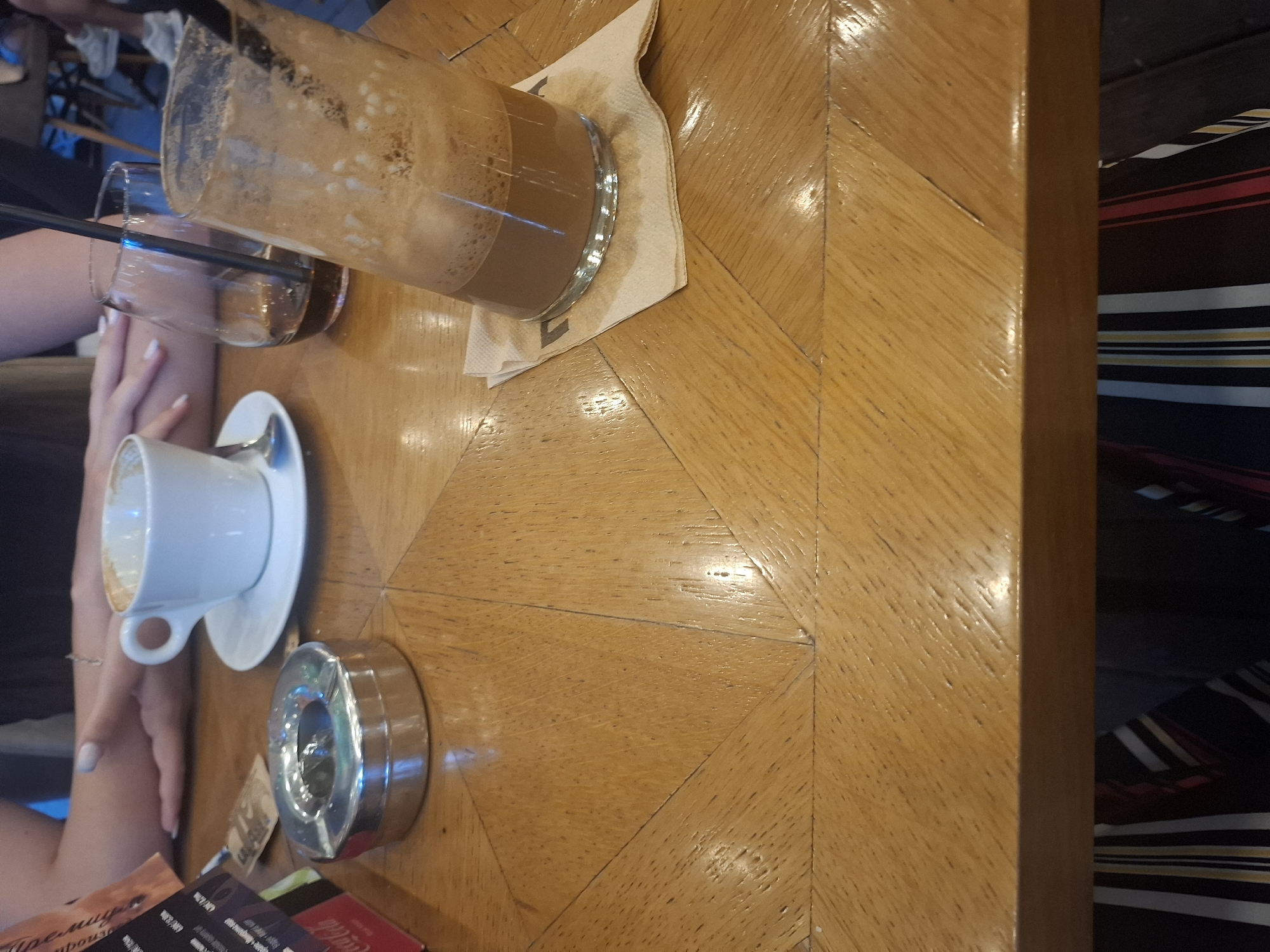 The image shows a close-up of a wooden table in what appears to be a cafe or restaurant. On the table are several items, including two glasses, one with a frothy brown beverage and a straw, and another partially filled with a dark liquid. A white teacup and saucer with a spoon are also visible, as is a shiny metal ashtray. The table has a patterned surface made of wood planks. In the background, a person's arm and hand can be seen, along with parts of other tables and chairs, suggesting an indoor public space. The lighting is dim, and the overall impression is of a casual setting. The image is oriented vertically. There is no visible text in the image.