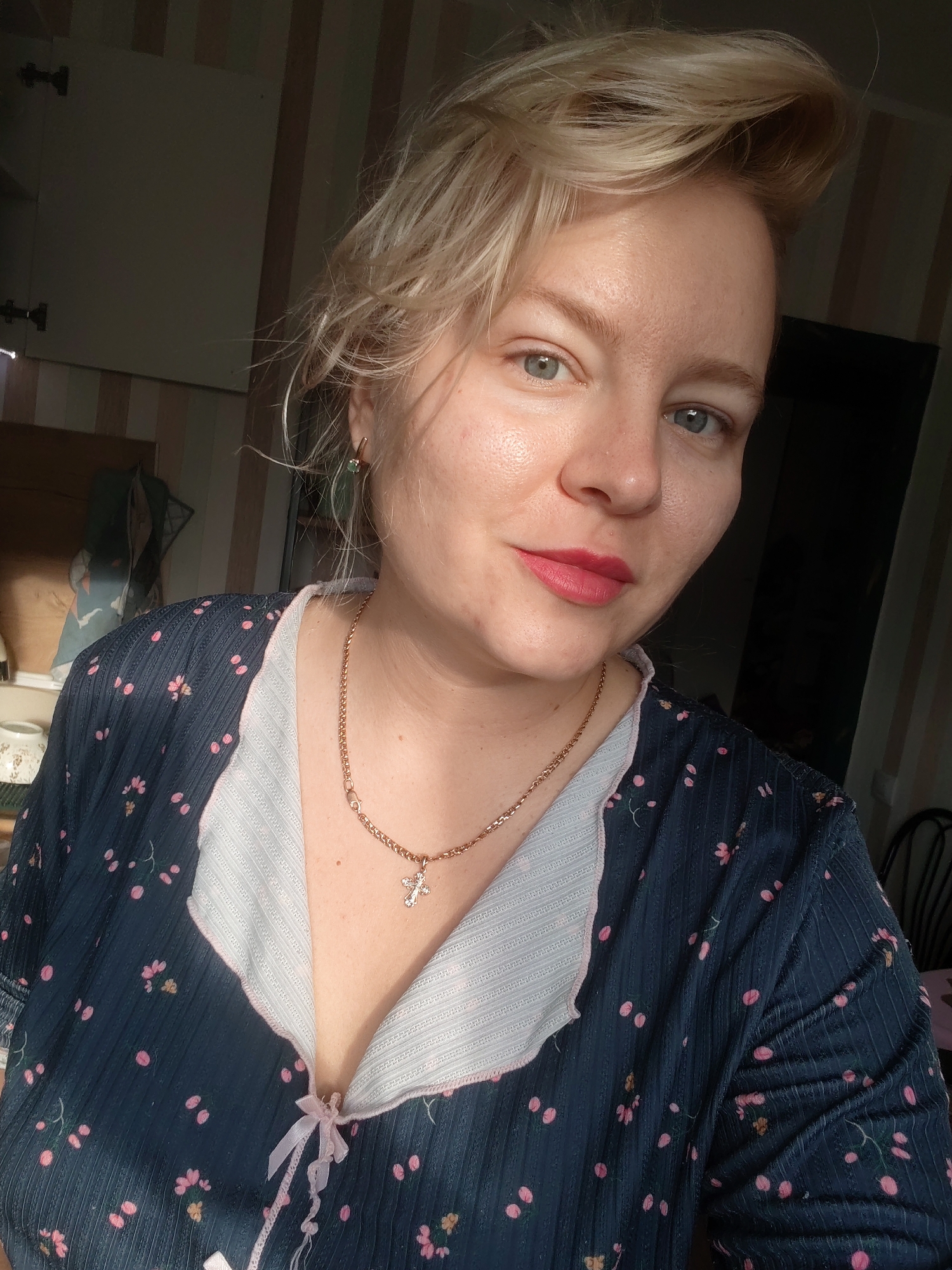 The image shows a close-up, waist-up, indoor portrait of a light-skinned woman with blonde hair, pale blue eyes, and pink lipstick. She is wearing a dark blue garment with a floral pattern of pink and yellow flowers. The garment has a light-colored trim with a pink edge and a pink bow at the neckline. She is also wearing a gold necklace with a cross pendant. The background appears to be a domestic setting, with striped wallpaper and what looks like the inside of a cabinet or closet visible to the left. The lighting suggests it is daytime, with sunlight casting shadows. The image does not provide any specific information about the location being Izhevsk, Russia.