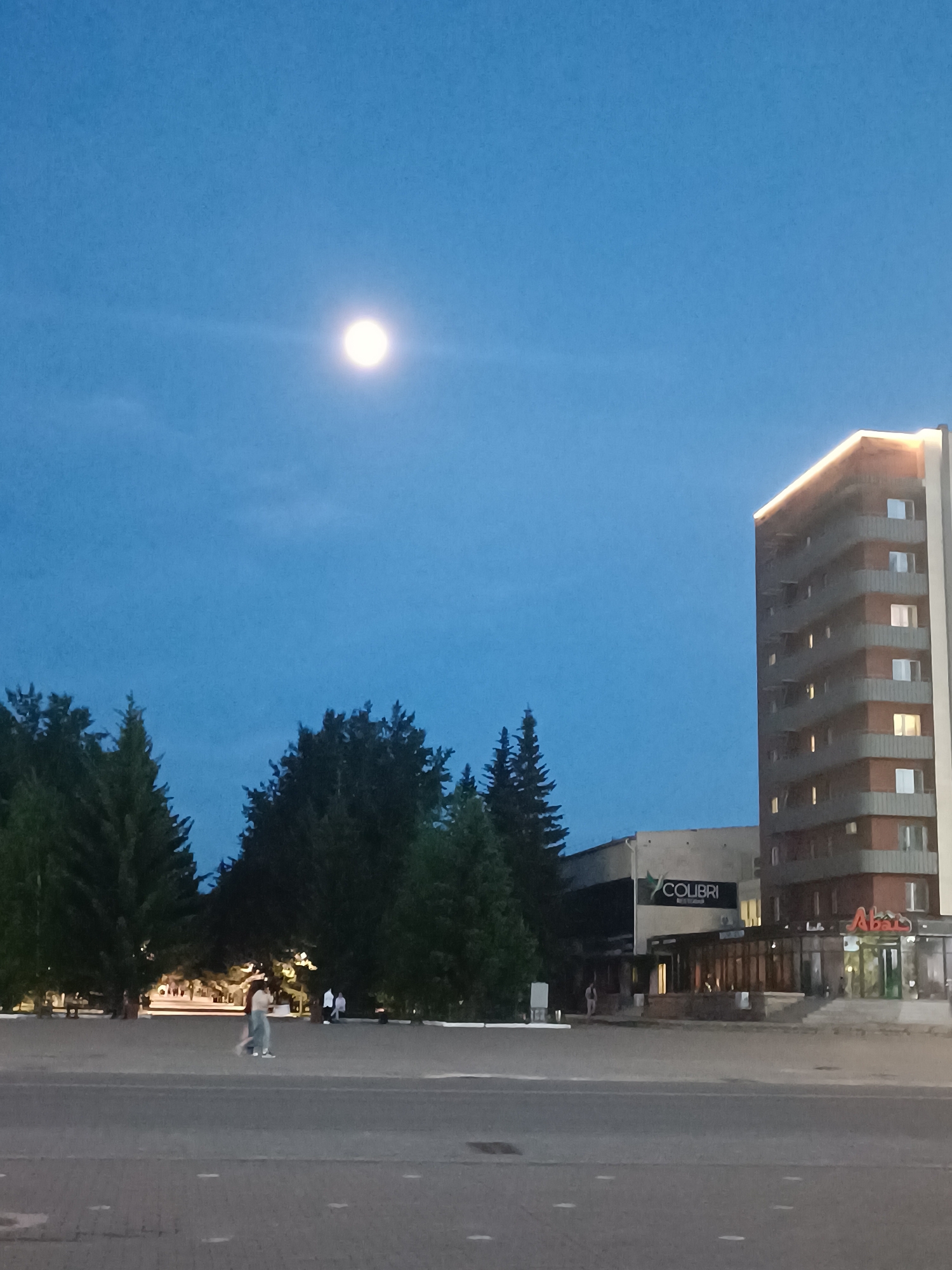 A nighttime scene captures a public square in Kokshetau, Kazakhstan, illuminated by a full moon in the upper sky. In the distance, a monument is visible, surrounded by trees and faint lighting. A few individuals are present near the monument. Adjacent to the square is a building complex featuring the "Colibri" restaurant and the "Abay" cafe. The restaurant is marked by its signage, while the cafe's name is illuminated. To the right of the building is a tall, multi-story structure with lit windows. The foreground is dominated by a paved open space, devoid of vehicles but traversed by a person walking across. The general ambience suggests a quiet evening in the city.