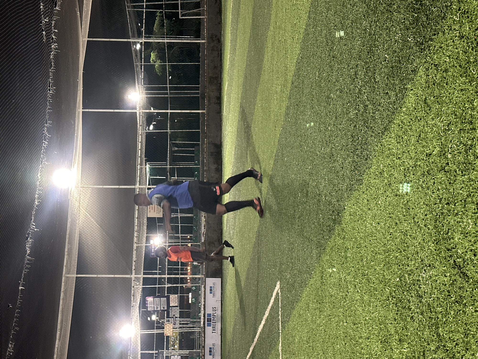 Two individuals are playing soccer on an artificial turf field under artificial lighting at night. The field is enclosed by a net and fencing. In the background, there are signs and trees visible. The scene appears to be at a sports facility in Abuja, Nigeria.