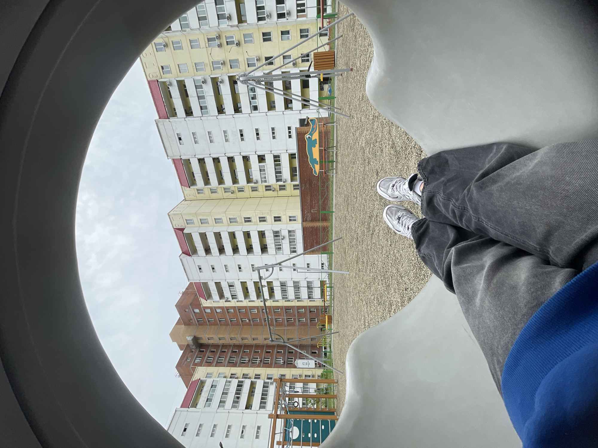 The image shows a low-angle view from inside a playground tunnel, with a person's crossed legs and white sneakers visible in the foreground. The playground is situated in front of a high-rise apartment building. The building has multiple floors, with a mix of yellow, white, and brown sections, and red-tiled roofs. The playground area has a tan gravel surface, and several play structures, including swings and climbing frames, are visible. The sky is overcast. The location appears to be Vladivostok, Russia, due to the architecture of the buildings and the general appearance of the environment.
