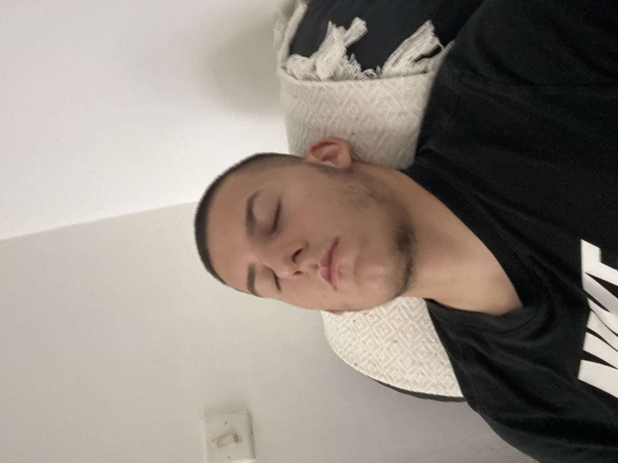 A man is lying down. He is wearing a black t-shirt with white text. His eyes are closed. He has short dark hair and stubble. His head is resting on a textured white pillow. A wall is visible in the background, and a light switch is on the wall. It is impossible to determine if this photo was taken in Burgas, Bulgaria.