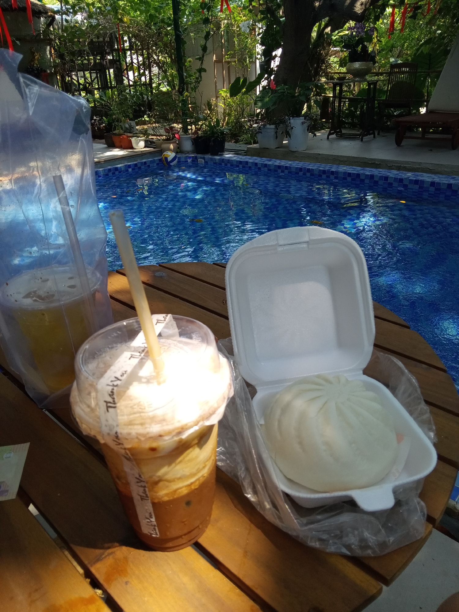 The scene depicts a still life arrangement set on a wooden surface, with a swimming pool visible in the background, possibly taken in the city of Bac Natural Heritage Area Trang, Vietnam.
On the wooden surface, there are two beverages: one is a yellow liquid in a plastic cup enclosed in a larger plastic bag, and the other appears to be an iced coffee drink with a white topping, also in a plastic cup with a decorative "Thank You" band around it. Beside the drinks, there is a white polystyrene food container opened, revealing a large, steamed bun inside, which is also contained within a plastic bag.
In the background, there is a swimming pool with blue and white tiles. Beyond the pool, there is vegetation, including trees and various potted plants. Further back, there are some outdoor chairs and a table, suggesting a patio or relaxation area. The overall environment suggests an outdoor setting, possibly a poolside area at a restaurant or resort.
