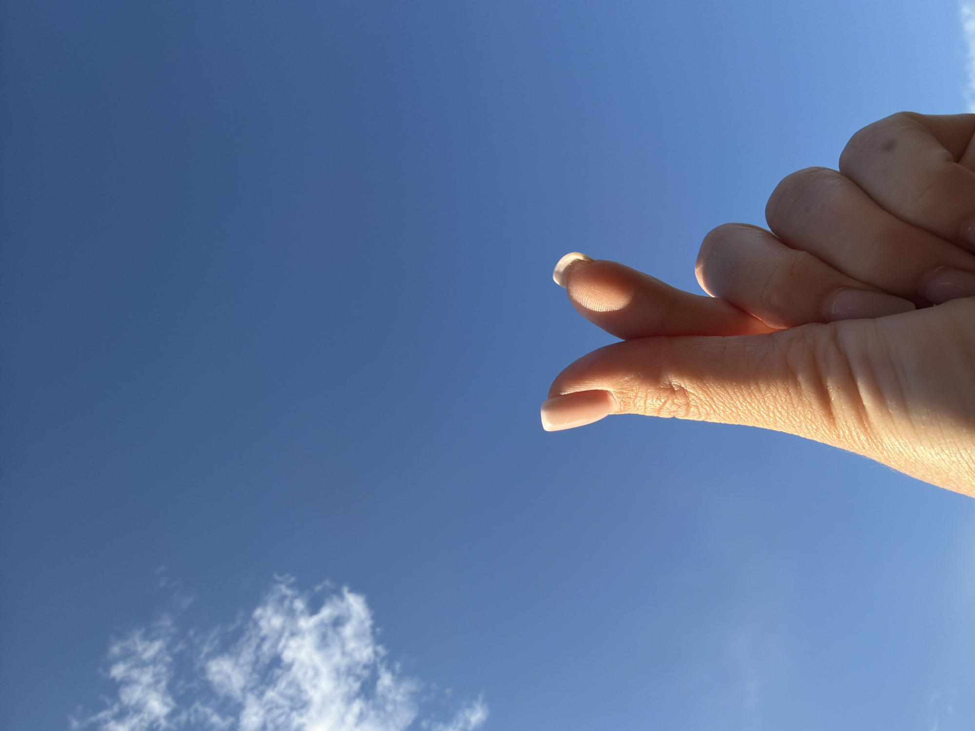 A close-up shot shows a person's hand forming a heart shape with their thumb and index finger against a clear blue sky with a few wispy clouds. The hand is positioned in the lower center of the frame, with the fingers extending upwards. The skin tone is light, and the fingernails are neatly manicured with a subtle sheen. The background is dominated by a vibrant blue sky, with a small cluster of white clouds on the left side of the image. The lighting suggests it is daytime, possibly late afternoon given the warm glow on the skin. The image does not contain any elements that place it within the city of Шмидтово, Russia.