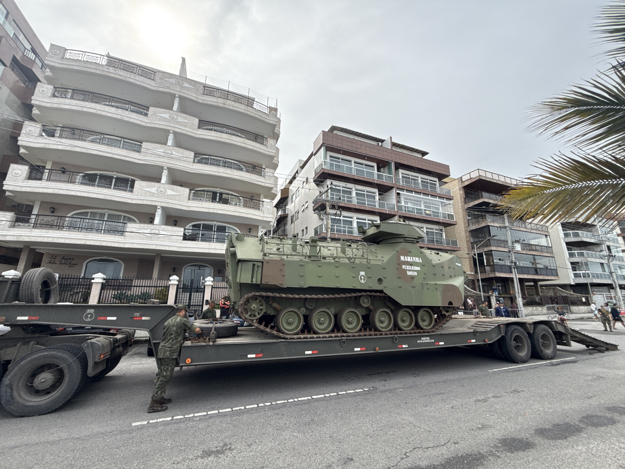 A military amphibious tracked vehicle, painted olive drab, is being transported on a flatbed trailer on a paved street in Cabo Frio, Brazil. The vehicle has the words "MARINHA FUZILEIROS NAVAIS" printed on its side, indicating it belongs to the Brazilian Marine Corps. Two individuals in military camouflage uniforms are visible near the trailer. One is standing by the trailer, seemingly adjusting something. The trailer is parked on the street, with its ramp extended. In the background, there are multi-story apartment buildings with balconies. A palm tree frond is visible in the upper right corner of the image. The sky is overcast.