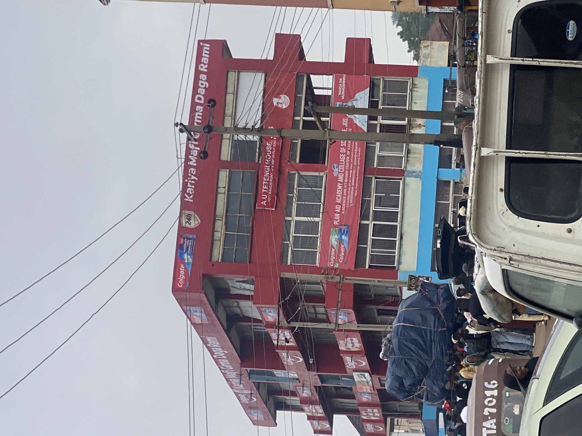 The image shows a multi-story red building in Jos, Nigeria, with various advertisements and signage. The building appears to be an office or commercial complex. On the top of the building, there is a large red sign with white lettering that says "Kariya Mafi Girma Daga Rami". Below this, there are several smaller signs, including "Colgate" advertisements and a sign for "A.U. TETENGI HOUSE, 4077 BAUCHI ROAD, JOS, NORTH PLATEAU S". Another banner on the building reads "PLAN AID ACADEMY AND COLLEGE OF SC...".
In front of the building, there is a street scene with several vehicles and a crowd of people. A large blue tarp-covered object is loaded onto a truck in the foreground. Several people are gathered around, some appearing to be waiting or directing traffic. The sky is overcast.