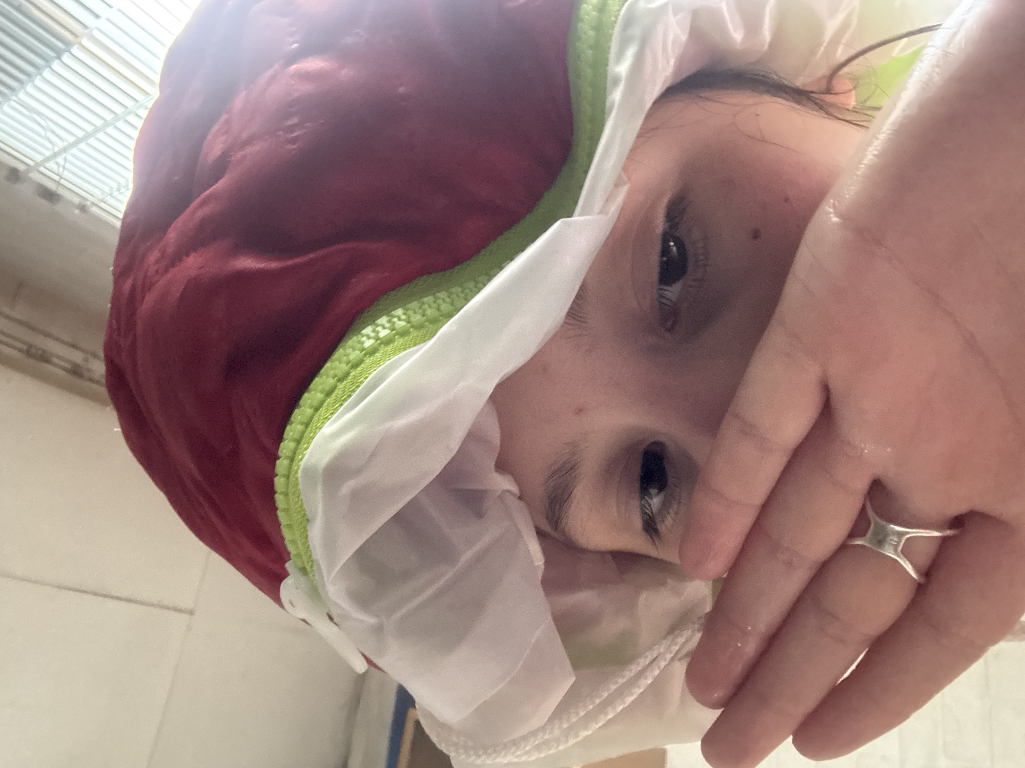 A close-up, high-angle photograph depicts a person wearing a red and white hooded jacket. The jacket has a green zipper. The person's face is partially obscured by a hand adorned with a silver ring. The location is not discernible from the image.