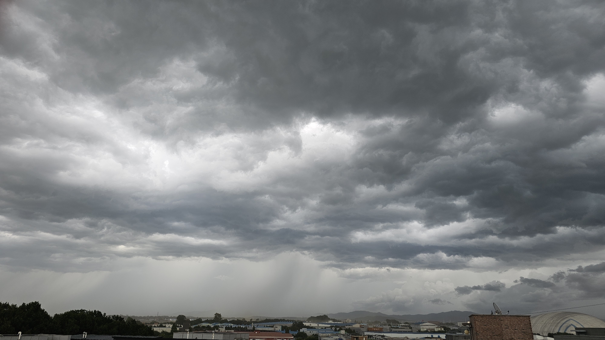 The image depicts a stormy sky over the city of Asan-si, South Korea. Dark, heavy clouds fill the upper portion of the frame, with streaks of rain visible in the distance over a low-lying urban area. The city appears to consist of industrial buildings and some residential structures, with trees lining the foreground. Mountains can be seen in the background. The overall atmosphere is one of impending or ongoing rainfall, characterized by a dim, overcast lighting condition.