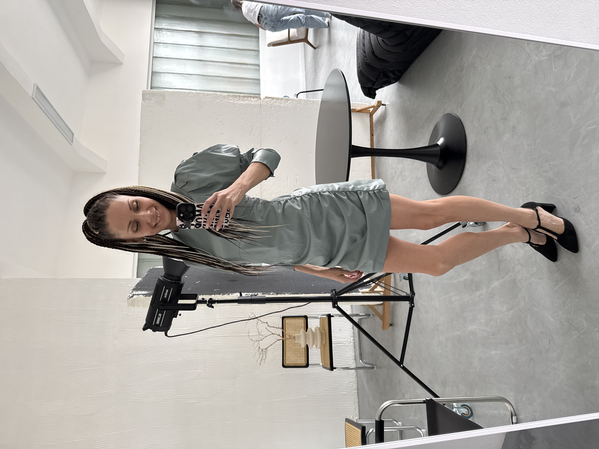 A young woman with braided hair stands facing a mirror, taking a selfie. She is wearing a light green dress and black Mary Jane style shoes. The background shows a photography studio setup with a studio light on a stand, a black table with a round top, and assorted chairs. The setting appears to be indoors with a concrete floor and white walls. The city of Bor, Russia is not evident in this image.