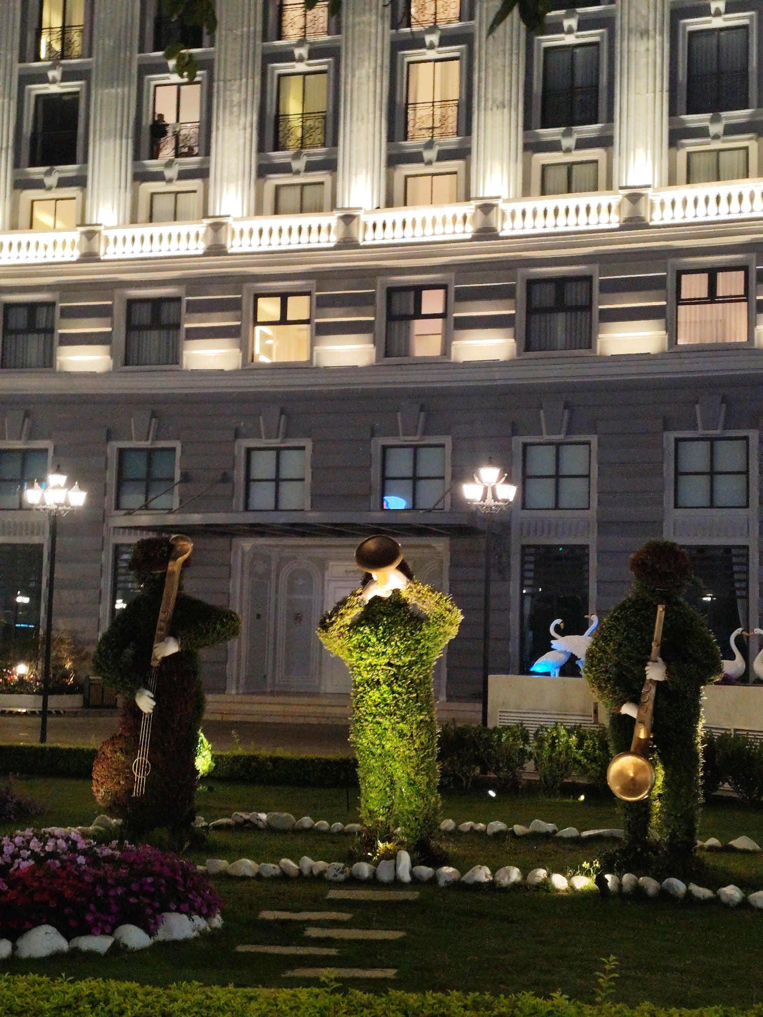 At night, the camera captures the formal gardens in front of the luxurious Palace Hotel Da Lat in Da Lat, Vietnam. The hotel, a multi-story building with windows and balconies, is lit with white lights. In the garden below, three topiary figures, shaped like musicians, stand amongst flowers and carefully laid stones. One holds a cello, another a trumpet, and the third a brass instrument. They are illuminated from below, creating highlights on their green foliage. In the background, swan sculptures sit in the garden. The overall scene suggests a curated and elegant aesthetic.