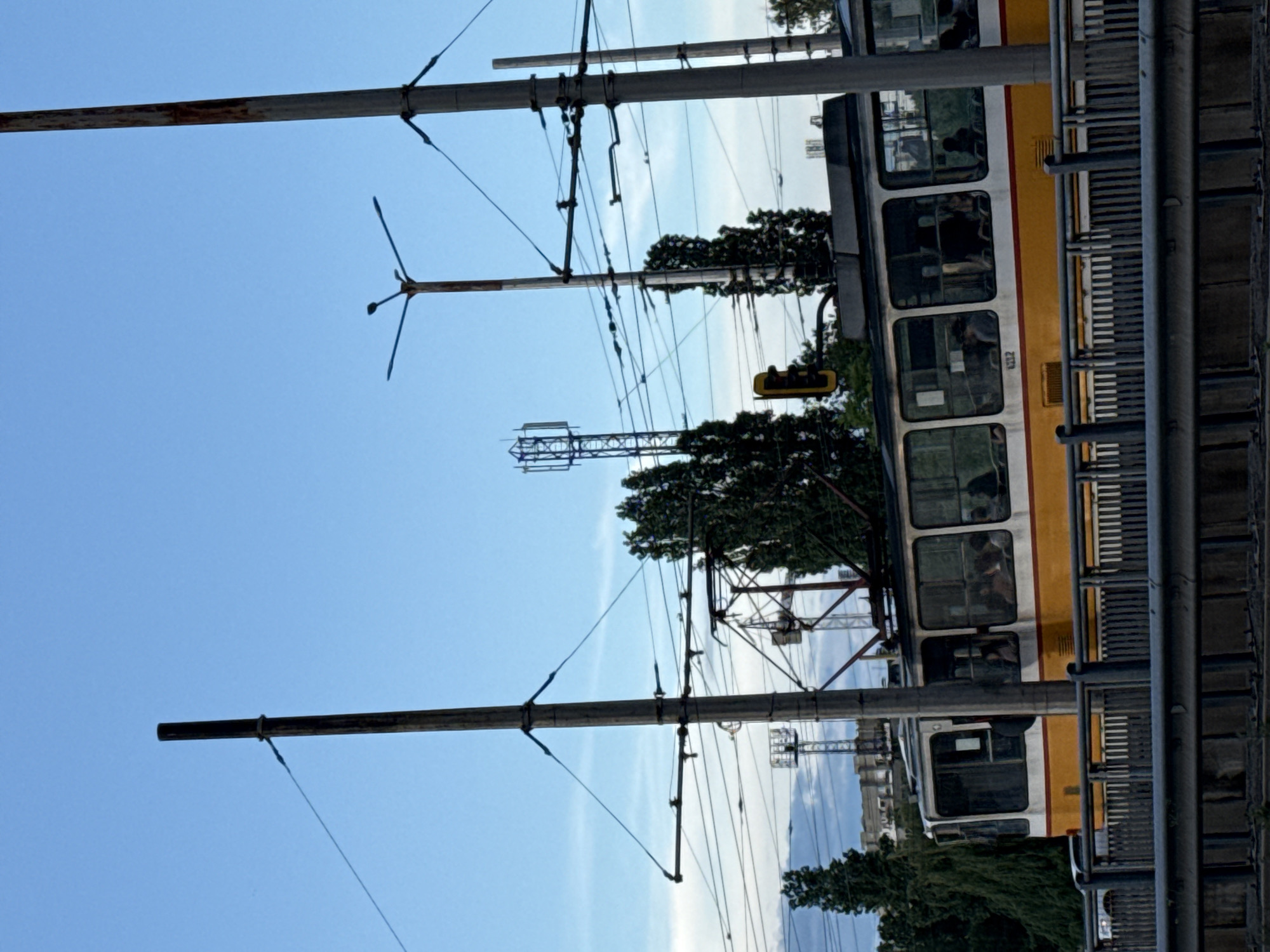 This daytime photograph captures a vibrant urban scene, likely in Sofia, Bulgaria, centered around a yellow and white tram. The tram, with a distinct yellow lower section and white upper body, is seen along its tracks, which are implied by the extensive overhead line system. Several passengers are visible as dark silhouettes through its windows, and the number "102" is faintly discernible on the side of the tram.
The upper portion of the image is dominated by a clear, bright blue sky, with a few faint wisps of white clouds visible in the distance. Spanning across this sky is a complex network of dark-colored poles and electrical wires, part of the catenary system that powers the tram. Numerous tall, slender poles with horizontal cross-arms rise from the ground, supporting these intricate overhead lines.
Green foliage from trees is scattered throughout the scene, notably a cluster on the right side that partially obscures a yellow traffic light or signal hanging from an overhead structure. A grey metal barrier or fence runs along the right edge of the frame, separating the tram's path from the viewer's perspective. In the far background, faint outlines of distant buildings can be seen against the horizon. The bright, even illumination suggests clear and fair weather during the day.