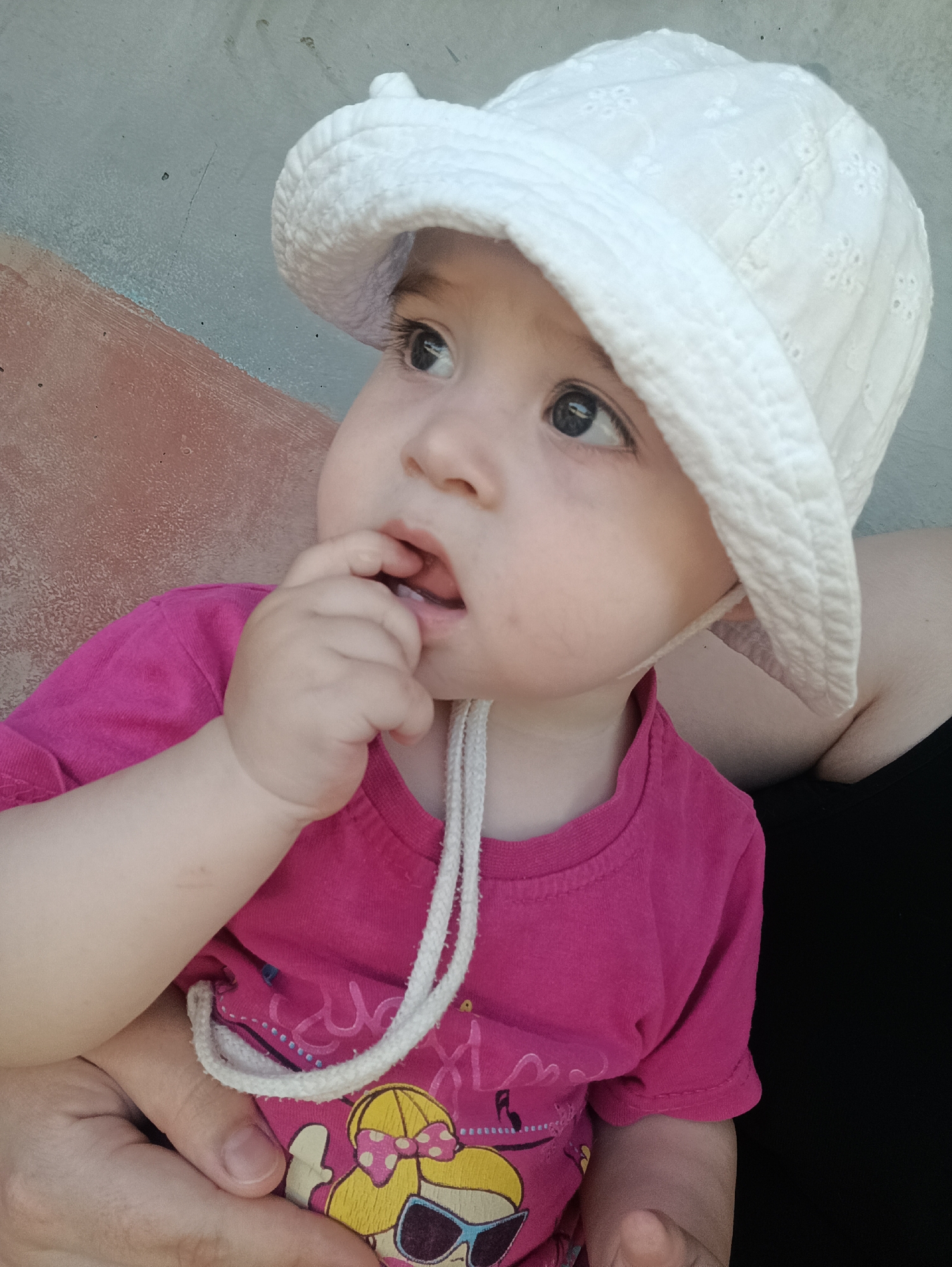A close-up shot shows a baby wearing a white sun hat and a bright pink t-shirt. The baby is looking to the left, with one hand in their mouth. The t-shirt has a graphic print of a cartoon character with blonde hair, sunglasses, and a pink bow. A white drawstring from the hat is visible, looped around the t-shirt. The background is a textured wall in muted tones of gray and reddish-brown. The lighting suggests it is daytime. The image appears to be taken outdoors, possibly in a casual setting like a backyard or park, given the natural light and textured background. The baby's expression is contemplative or perhaps a bit fussy. The location context is Посад, Russia.