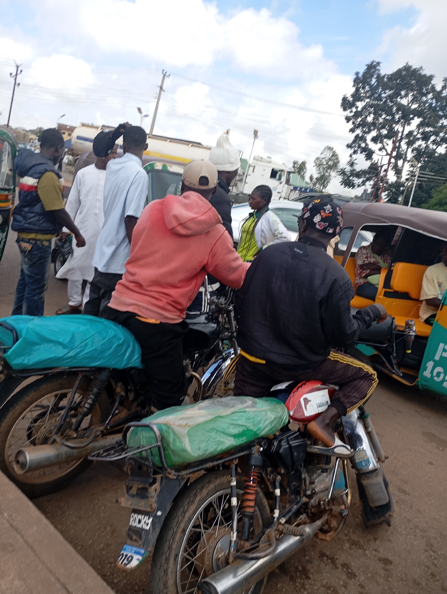 The image captures a street scene in Gura Top, Nigeria, with multiple people and vehicles present. In the foreground, two motorcycles are parked. The motorcycle on the left is partially covered with a teal tarp, and the motorcycle on the right has a green tarp covering its rear seat. A person wearing a black jacket and striped pants is seated on the right motorcycle, facing away from the camera. A person in a pink hooded sweatshirt is seated on the left motorcycle, also facing away. Several other individuals are standing or walking in the background. To the right of the motorcycles, a three-wheeled auto-rickshaw with yellow seating is visible, carrying passengers. In the far background, a large truck and other vehicles are present, along with utility poles and trees under a cloudy sky. The ground appears to be a dirt or gravel road.