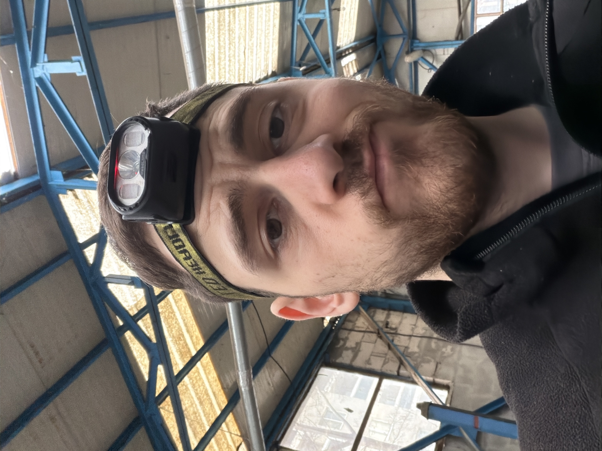 The image shows a selfie of a man with brown hair and a beard, wearing a black headlamp with a green strap. He's wearing a dark-colored fleece jacket or sweatshirt. The background appears to be an industrial or warehouse setting with a metal-beamed ceiling painted blue. There are also windows visible. It is not possible to ascertain the precise location in Plovdiv, Bulgaria from the image.