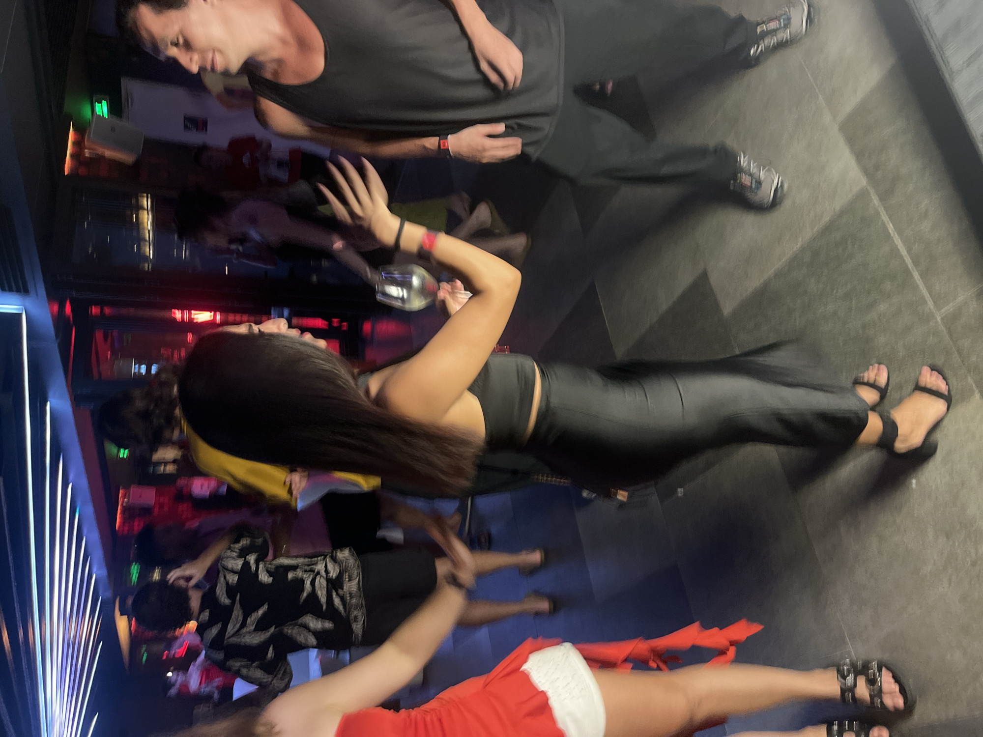 The image depicts a dimly lit, indoor social gathering, likely a nightclub or bar, in Đà Nẵng, Vietnam. The environment is characterized by ambient lighting from overhead neon-like fixtures and some red illumination in the background. The floor is tiled with large, dark gray tiles.
In the foreground and mid-ground, several individuals are engaged in social interaction, appearing to be dancing or socializing.
On the left, a woman in a red dress with white trim is partially visible from the chest down. She is wearing black sandals with multiple buckles.
To her right, a woman with long, dark hair is wearing a black crop top and black high-waisted pants. She is holding a glass of white wine in her left hand and her right arm is extended, with her palm open. She is wearing a red bracelet on her right wrist.
To the right of the woman in black, a man is visible from the chest down, wearing a black tank top and dark pants. He is wearing athletic shoes. He has a red bracelet on his left wrist.
In the background, other individuals are present but less distinct due to motion blur and lighting. One person is wearing a black shirt with a white pattern, and another is wearing a yellow shirt. The overall scene suggests a lively, nighttime event.
