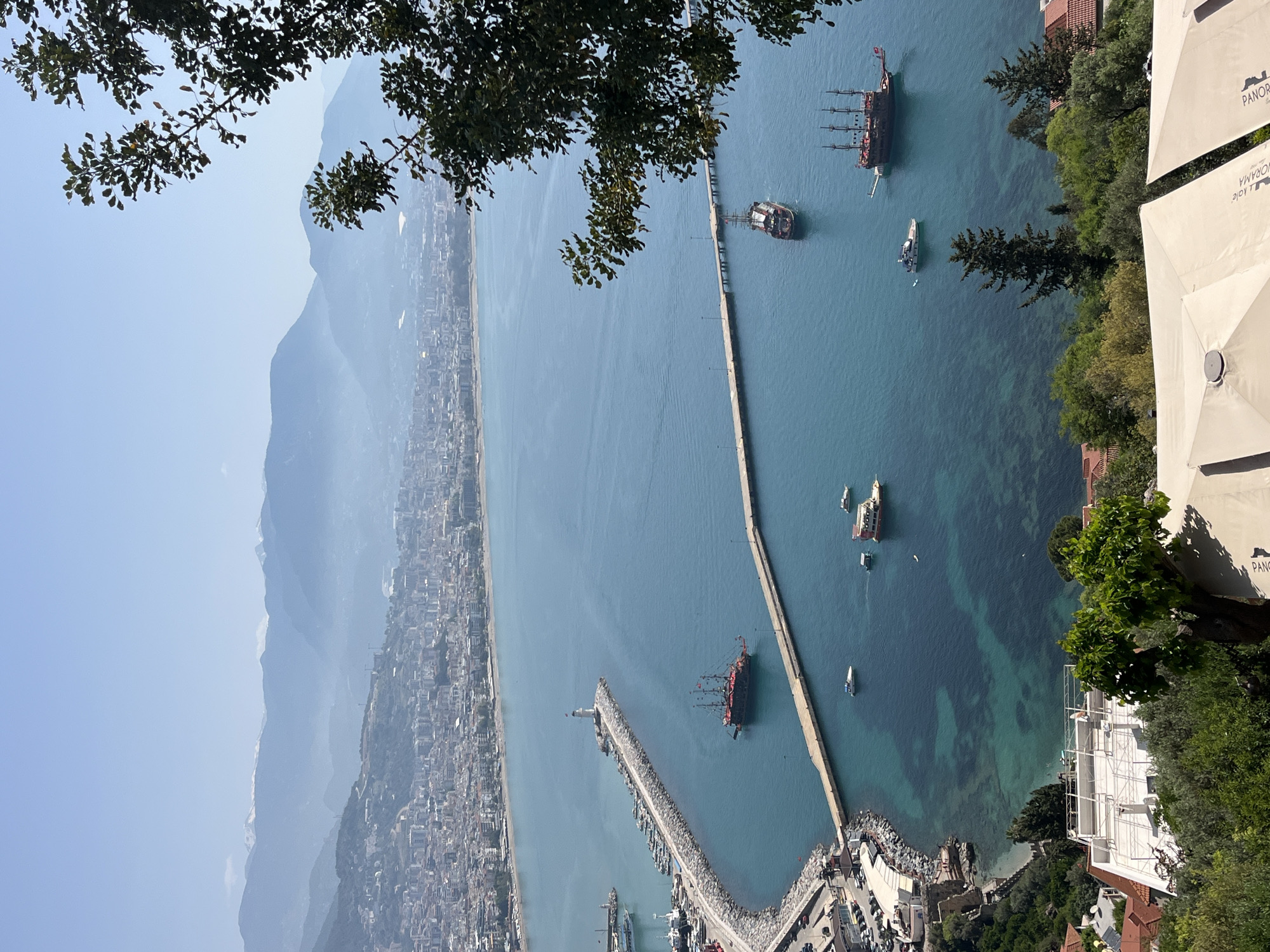 The image presents a panoramic view of Alanya, Turkey, featuring the city skyline, mountains in the background, and a prominent harbor. A long pier extends into the turquoise water, with several boats docked or anchored nearby, including a distinctive pirate ship. The camera's elevated perspective is partially framed by foliage in the foreground. The lighting is bright, suggesting a sunny day.