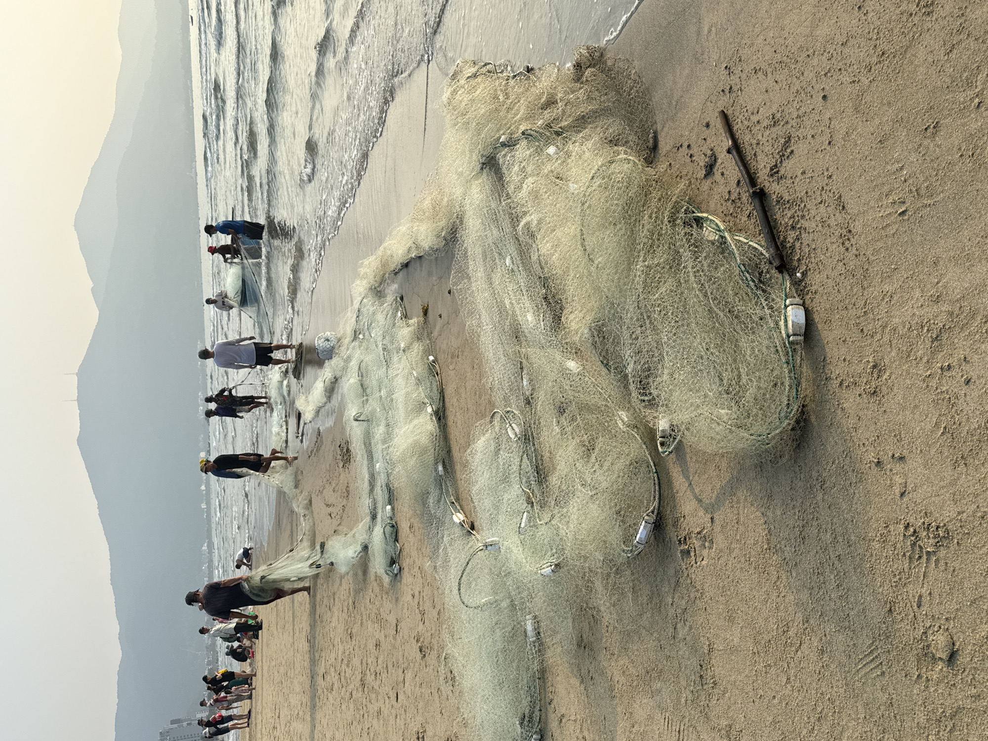 The scene is a beach in Đà Nẵng, Vietnam. A large fishing net lies on the sand, extending from the shoreline towards the right. Several people are pulling the fishing net. The beach transitions from the shoreline with gentle waves to a sandy expanse. In the background, mountains are visible through the haze. A dark, stick-like object rests near the fishing net on the sand. The fishing net is white. The sand is light brown.