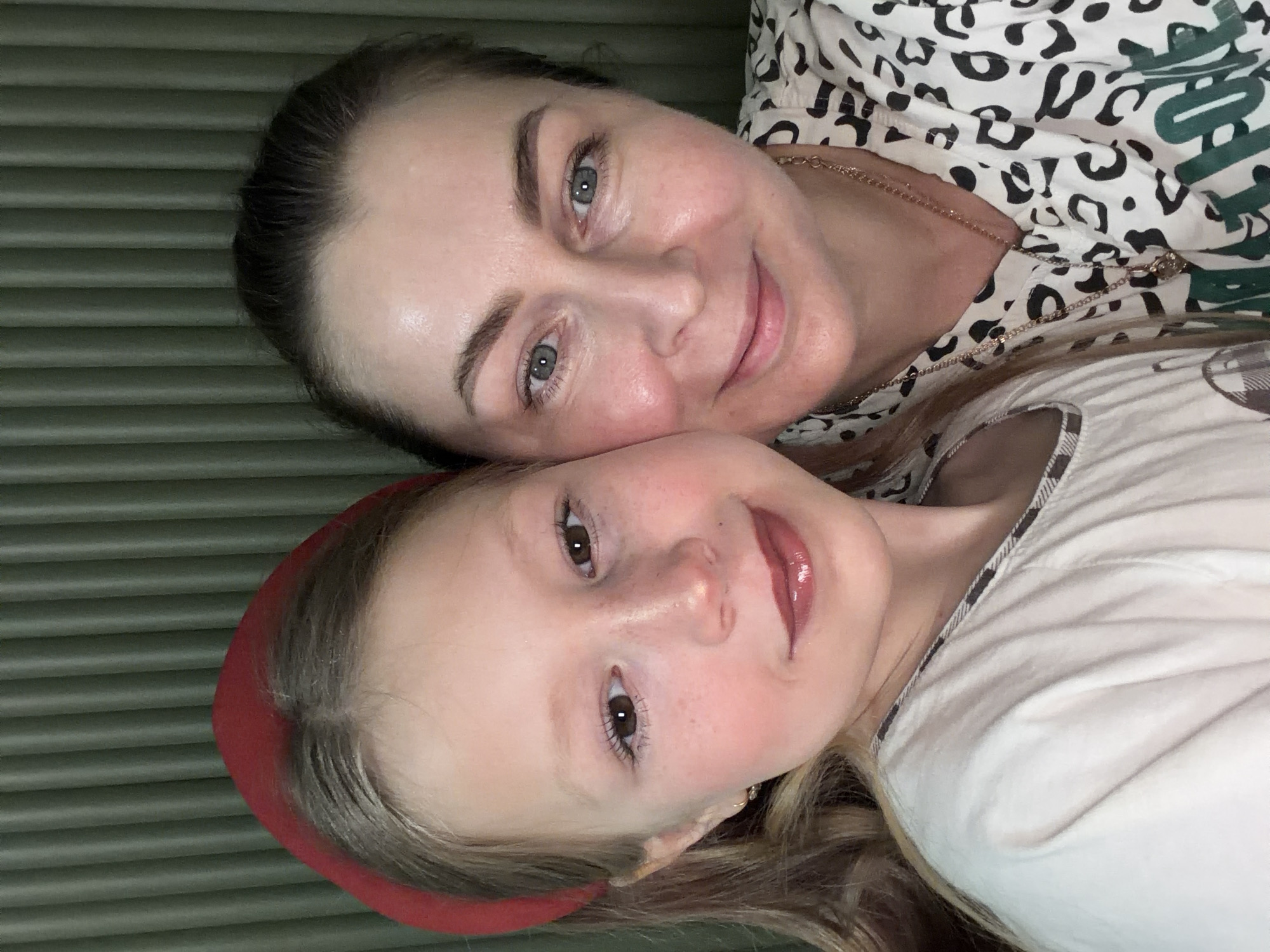 This close-up portrait features two females, an adult woman and a younger girl, likely a mother and daughter, positioned closely together in an indoor setting. Both subjects are looking directly at the camera with gentle, contented smiles, conveying warmth and affection.
The adult woman, on the upper left, has dark hair pulled back and light-colored eyes. She is wearing a patterned top featuring black and white animal print with accents of green, and a delicate gold chain necklace. The younger girl, on the lower right, has lighter-colored hair, dark eyes, and is wearing a vibrant red headband and a white top, possibly with a subtle checkered pattern. Her lips are glossy.
The background is a consistent muted olive-green color with horizontal ridged or slatted textures, suggesting a wall, headboard, or piece of furniture. The lighting appears soft and even, typical of an indoor environment. No specific time of day or weather conditions are discernible from the image. No visible text is present. The provided location context is Dubovaya, Russia.