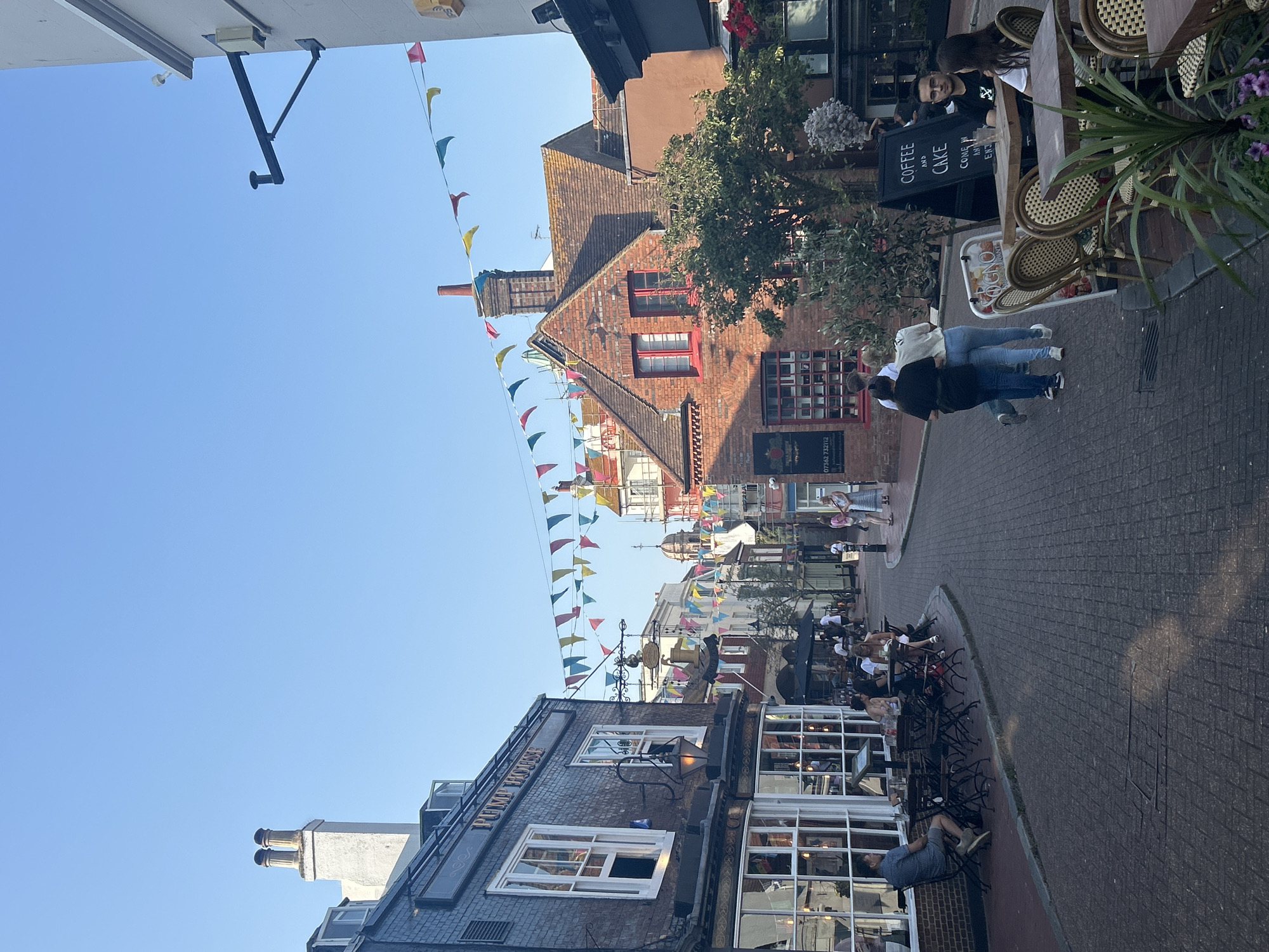 The image depicts a street scene in Brighton, United Kingdom, likely an area with pedestrian access and outdoor seating for establishments. The street is paved with cobblestones. String pennant flags in various colors are stretched across the street above the buildings.
On the left, a dark brick building with large white-framed windows and the sign "PUMP HOUSE" is visible. Several people are seated at tables outside this establishment. Further down the street, the buildings are a mix of brick and white stucco, with some sporting decorative architectural elements and chimneys. Red-framed windows are noticeable on a prominent brick building.
On the right side of the street, there is an outdoor seating area with tables and chairs, some occupied by diners. A blackboard sign reads "COFFEE AND CAKE COME AND ENJOY". A tree with green foliage is positioned between the brick building and the outdoor seating area.
In the foreground, people are walking along the street. Two individuals are walking away from the camera, one in a black top and blue jeans, the other in a white top and light blue jeans. Further on, a couple is seated at a table, with a man in a black t-shirt and a woman in a white top.
The sky is clear and blue, indicating daylight. The lighting suggests it is either early morning or late afternoon due to the long shadows cast on the cobblestone street. The overall atmosphere appears relaxed and commercial, typical of a busy street in a seaside town.