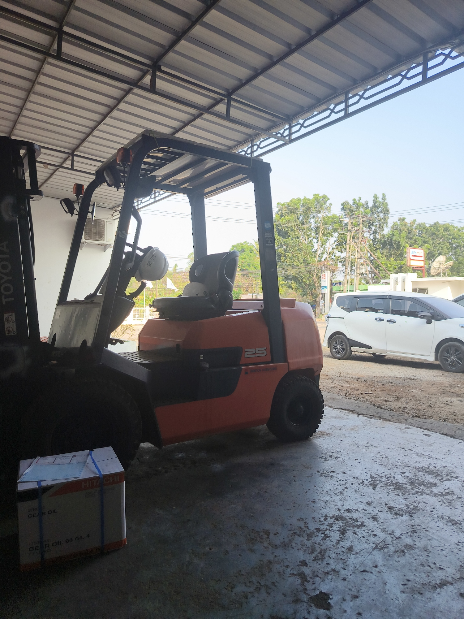 A Toyota forklift is parked under a metal awning. The forklift is orange and black, with the number "25" visible on its side. In front of the forklift, there is a cardboard box with blue straps and text that includes "HITACHI" and "GEAR OIL". To the right of the forklift, a white minivan is parked outdoors. The scene appears to be in an industrial or commercial area, possibly a warehouse or a lot, with trees and utility poles visible in the background under a clear sky. The ground is concrete and somewhat worn. The image is taken from a low angle, looking up towards the awning and the sky. There are no people visible in the image. The context suggests a scene in Pangkalpinang, Indonesia, likely an outdoor storage or operational area.