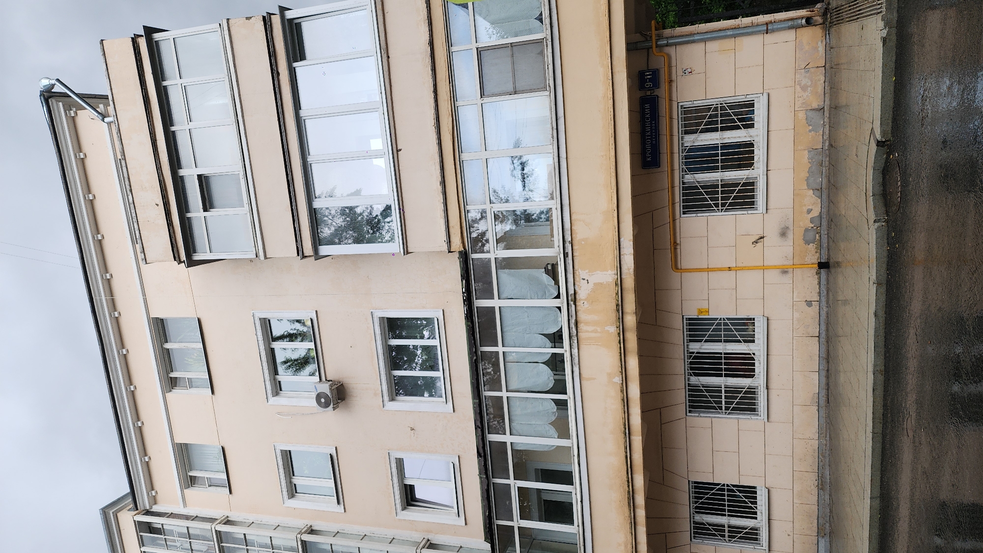 Here's a description of the image:
The photograph shows a building facade in Moscow, Russia. The building appears to be multi-story with many rectangular windows, some of which are barred with metal grates. A sign on the building reads "Кропоткинский переулок 9-11," indicating that the building is located on Kropotkinsky Lane and has an address of 9-11. A yellow gas pipe runs along the exterior wall. The adjacent sidewalk is wet, suggesting recent rain. The building's architecture has a Soviet-era aesthetic.