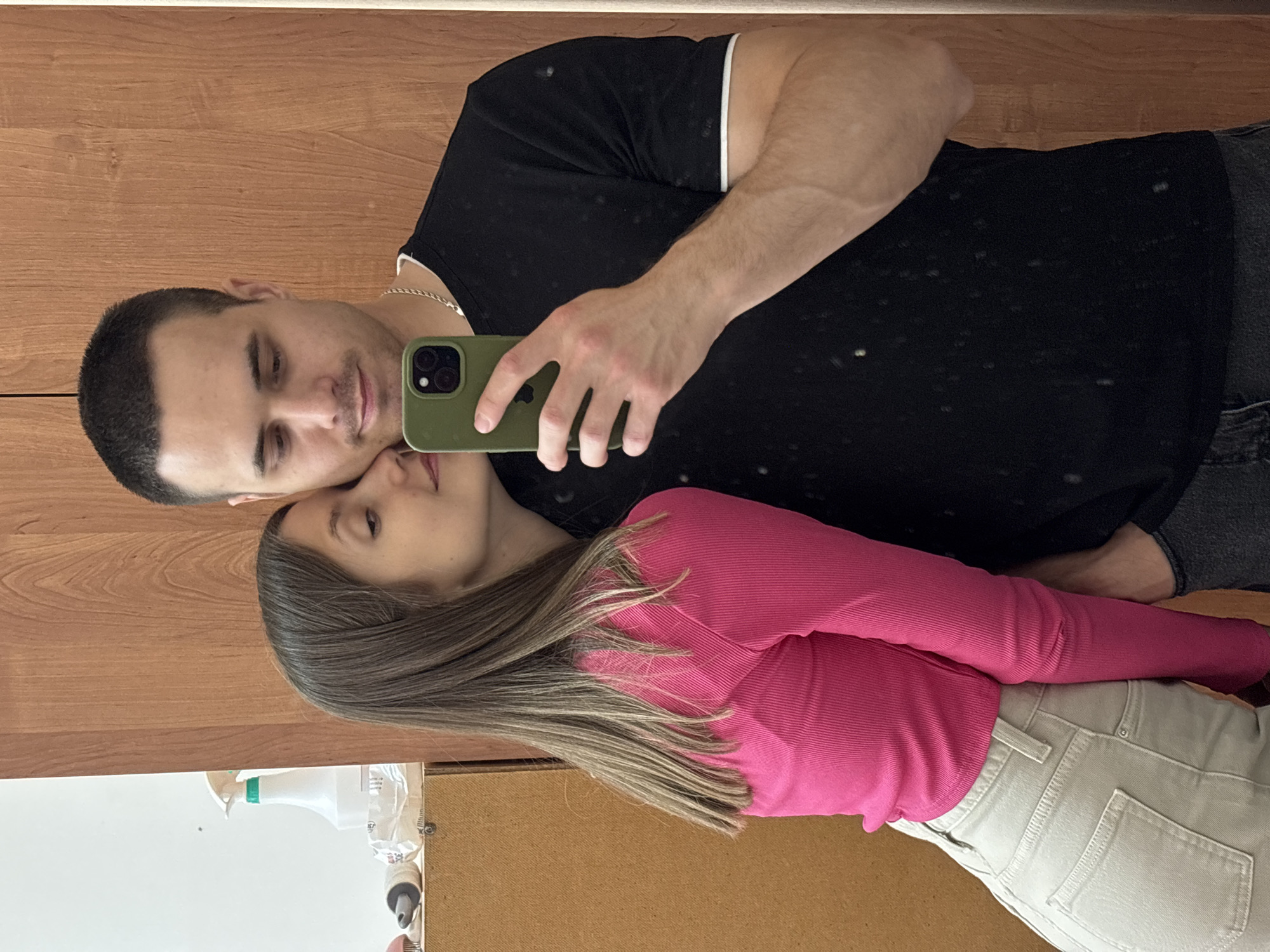An indoor medium shot shows a young man and woman posing for a selfie taken in a mirror. The man, positioned on the right, is looking down at his phone, which he holds in his right hand. He has short dark hair and is wearing a black t-shirt with a white trim around the collar and sleeves, and a silver chain. The woman is on the left, leaning her head against the man's shoulder and looking towards him. She has long brown hair and is wearing a bright pink long-sleeved top and light-colored jeans. The background consists of a wooden cabinet on the left and a wooden paneled wall on the right. A white spray bottle and other items are visible on the cabinet. The overall setting suggests a residential or personal space, possibly within the city of Chaikovsky, Russia, based on the provided context.