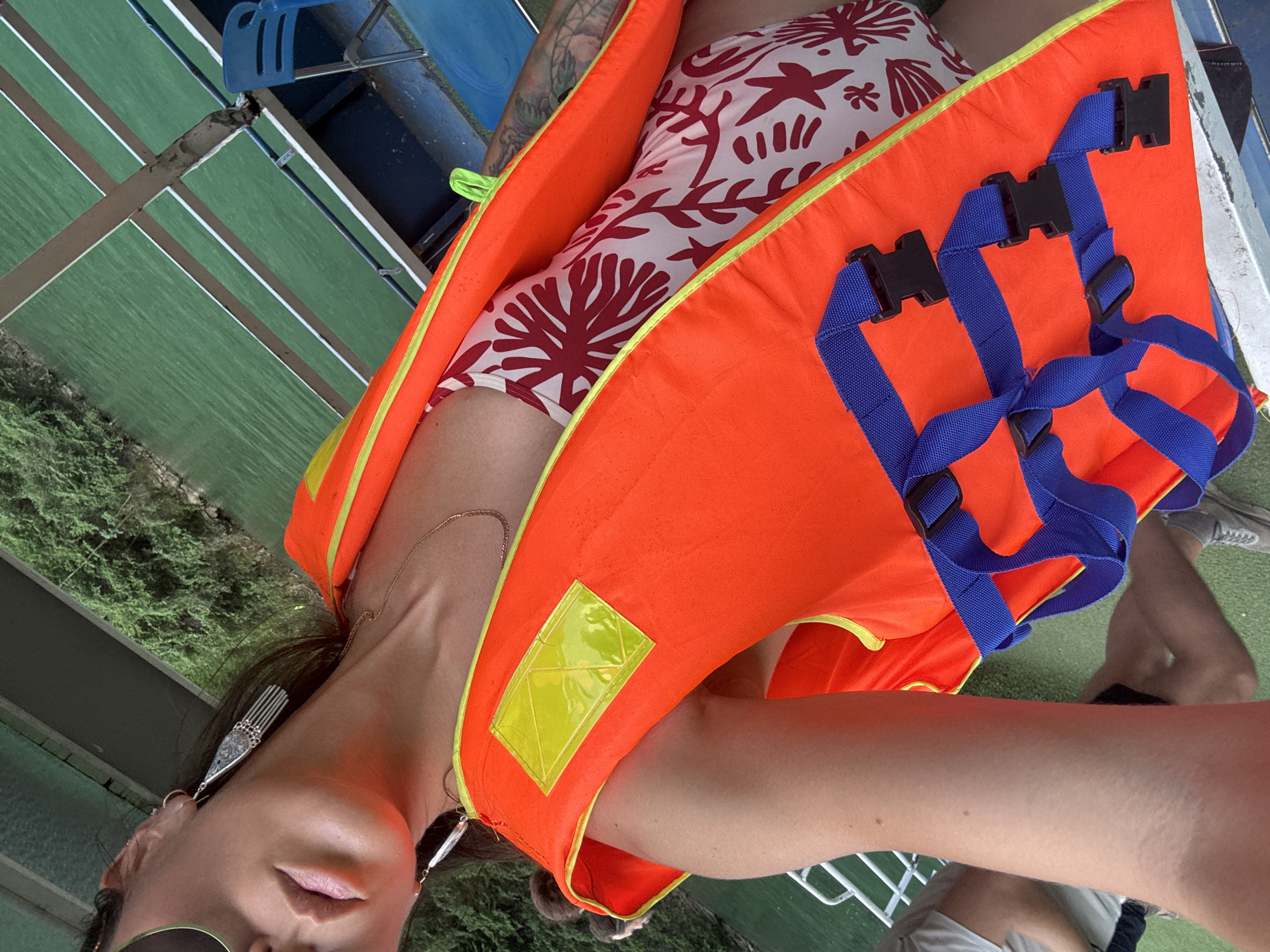 A woman, possibly on a boat in Đồng Văn Commune, Vietnam, is wearing a life vest. The vest is bright orange with blue straps and black buckles. She wears a white bathing suit with a red pattern, long silver earrings, and sunglasses. The boat's interior has green and white-striped surfaces, and the surrounding environment includes green foliage and water. Another person's legs and shoes can be seen partially in the bottom right corner.