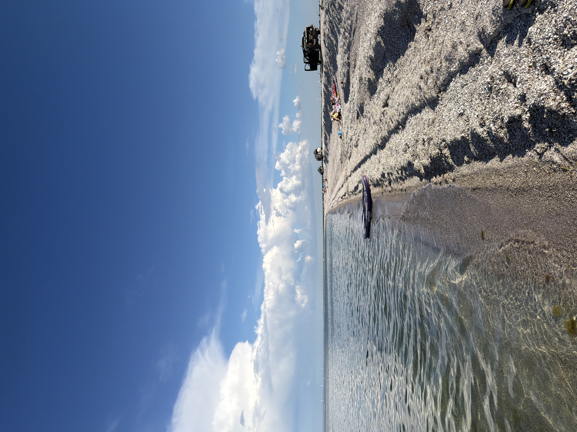 The image depicts a wide, expansive beach composed of crushed shells under a bright blue sky dotted with fluffy white clouds. The calm, shallow water gently laps at the shore, its surface rippled and reflecting sunlight. On the beach, several people are visible. Some are sunbathing on towels, while others are near parked vehicles. Two dark-colored SUVs and a pickup truck are parked on the sandy bank, suggesting a recreational outing. A purple inflatable paddleboard rests near the water's edge. In the distance, several wind turbines are visible on the horizon, indicating a coastal or open landscape. The overall scene conveys a sense of leisure and outdoor activity on a sunny day.