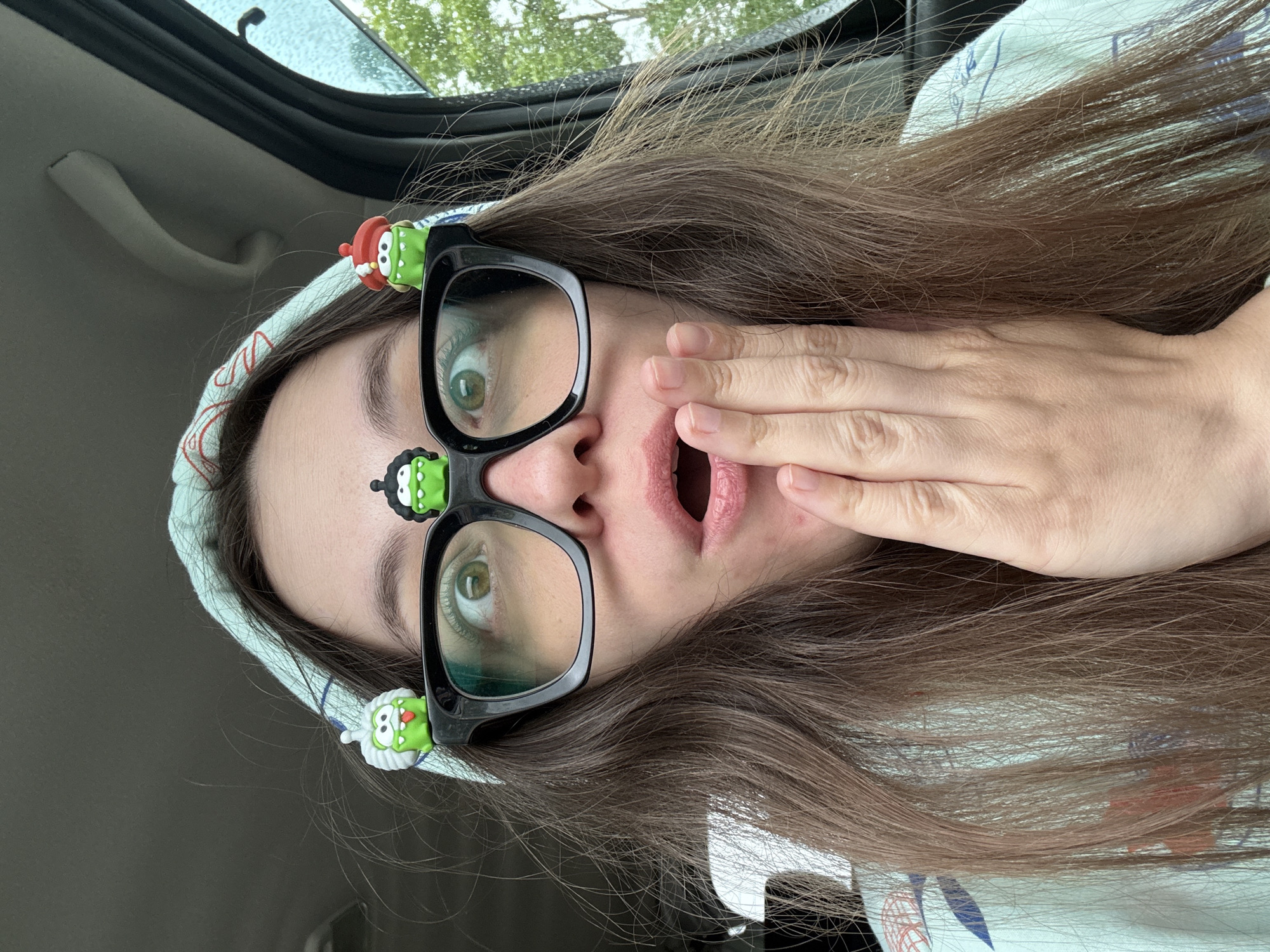 A woman with long brown hair is seated in a car. She is wearing large black-framed glasses adorned with cartoonish green and white figures, and a blue and white patterned headscarf. Her mouth is open, and her hand is partially covering her mouth and cheek. The background shows the interior of a car, including the door frame and window. The location in the city of Фоки, Russia is not discernible from the image.