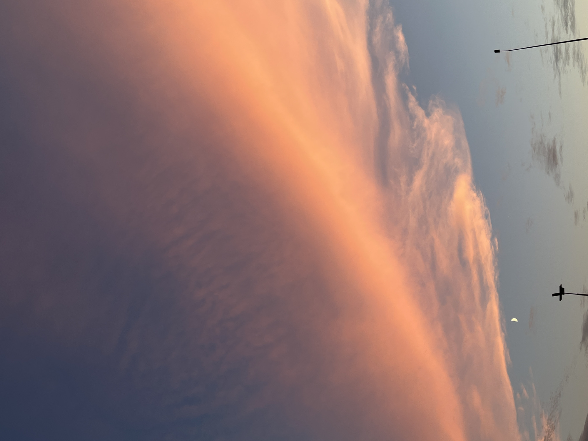 This is a photograph of a twilight sky over Saint Petersburg, Russia. A large band of clouds stretches diagonally across the frame from the lower left to the upper right. These clouds are illuminated from the side by the setting sun, casting a warm, peachy-orange glow. The upper portion of the sky is a deep, dusky blue, transitioning to a lighter blue towards the horizon. A sliver of the moon is visible in the lower left quadrant of the image, just above a dark, silhouetted structure that appears to be a lamppost or street light. Another similar silhouetted lamppost is visible on the right edge of the frame, further in the distance. The overall impression is one of a serene and atmospheric sunset scene in an urban environment.