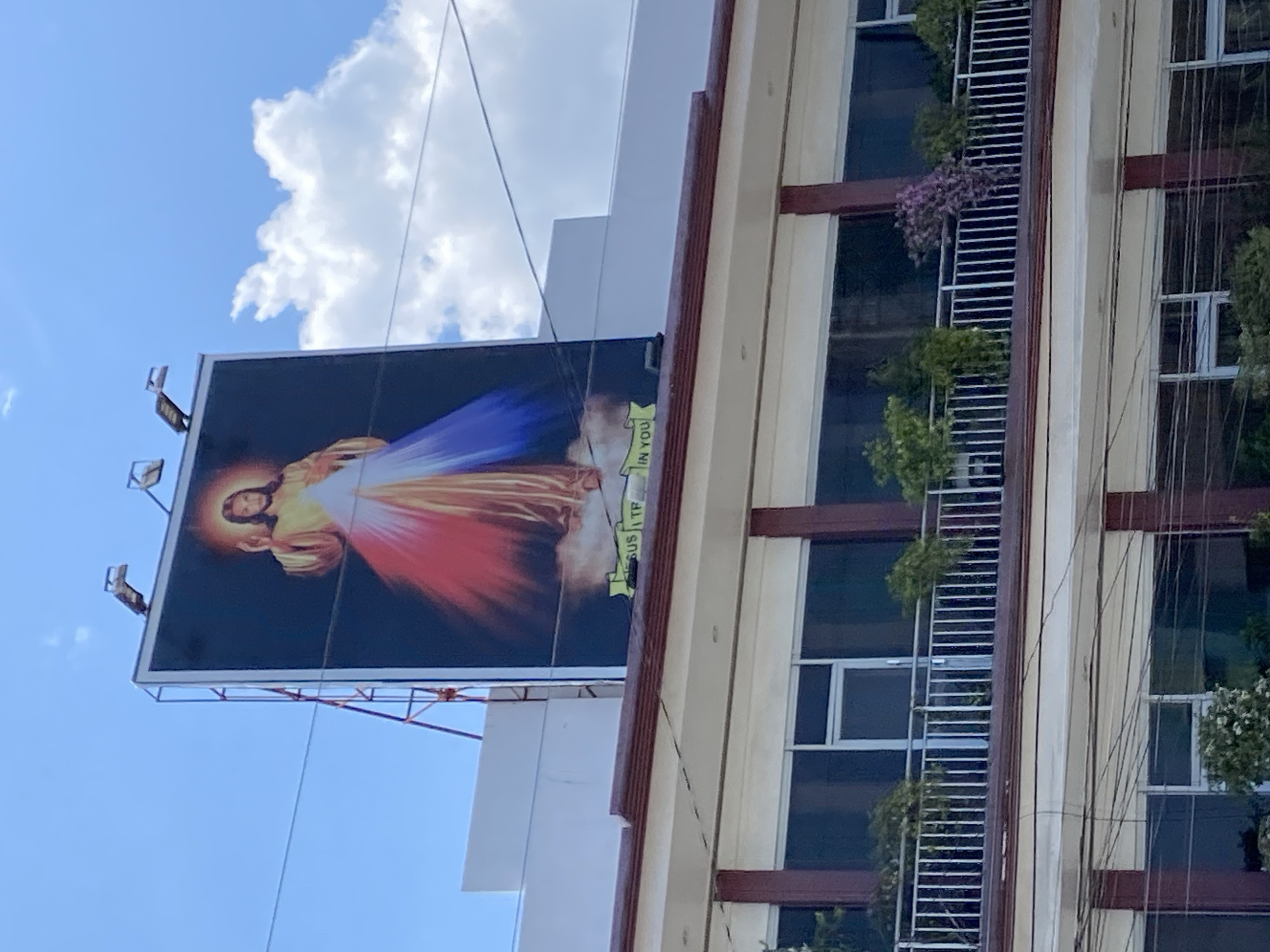 The media file, presented rotated 90 degrees counter-clockwise, captures an outdoor urban scene in Davao City, Philippines, under a bright blue sky with scattered white clouds.
The primary subject is a large billboard displaying a religious image of Jesus Christ, known as the Divine Mercy. Jesus is depicted with one hand raised in blessing and the other pointing downwards, from which emanate prominent rays of red and blue/white light. A green banner with yellow text is visible along the lower edge of the billboard in its current orientation, showing the words "IN YOU." The full text is understood to be "JESUS I TRUST IN YOU." The billboard structure includes a metal frame and several spotlights attached to its top edge.
Adjacent to the billboard, occupying the right side of the image, is a multi-story building. This building features numerous dark-paned windows set within white frames and several balconies or ledges. These balconies are adorned with lush green plants, and a vibrant cluster of purple flowers is visible on one balcony in the mid-section of the building. Utility wires or cables run vertically along the building's facade. The scene suggests a bright, sunny daytime environment.