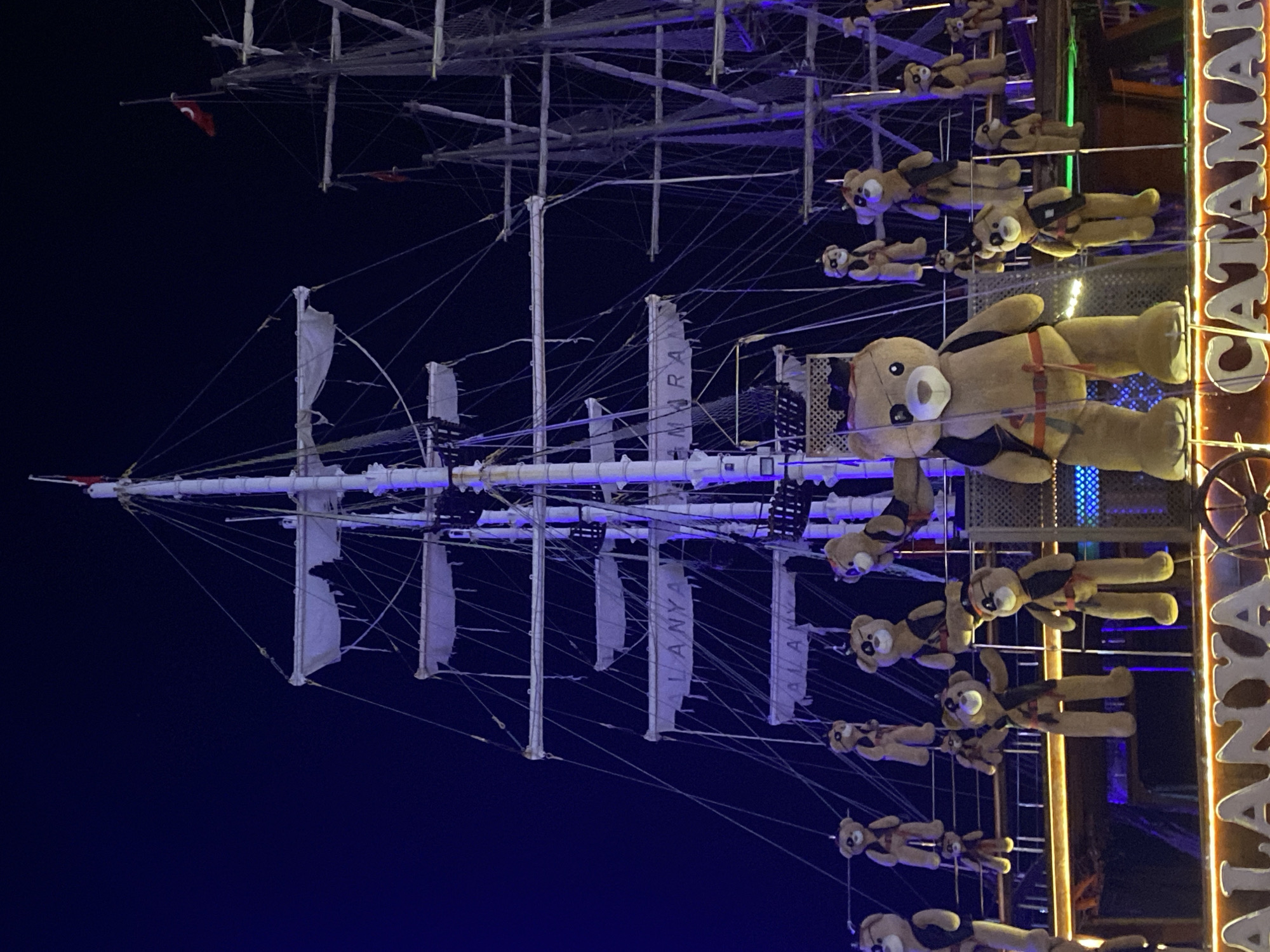 The image depicts an illuminated replica of a sailing ship, with masts, rigging, and partially furled sails. The word "ALANYA" is displayed vertically on the sails. To the right of the ship model is a vertical sign that reads "CATAMARAN." Teddy bears wearing harnesses are suspended from the ship's structure, appearing to be in freefall. The background is a dark blue night sky. The location is likely in Alanya, Turkey, given the name on the sails. The scene is brightly lit, and the sign suggests a tourist attraction or business related to catamaran tours.