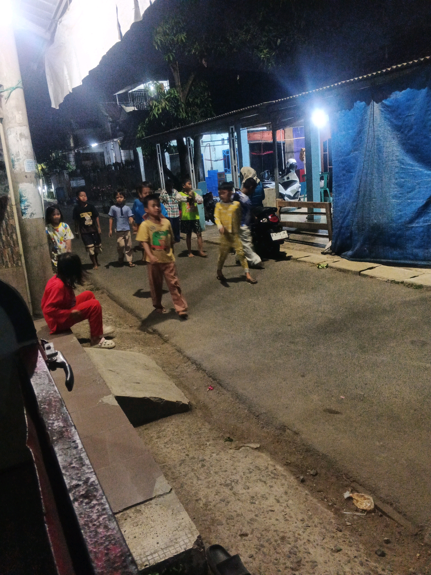 This is a nighttime, eye-level, full shot of a street in Kadu, Indonesia, with several children present. The scene is lit by artificial lights, creating shadows and a slightly grainy effect. A group of children, estimated to be between five and ten years old, are dispersed across the paved street and sidewalk area.
In the foreground on the left, a child in a red shirt and pants is seated on a concrete ledge. To the right of this child, the street is visible, with a few scattered debris items including a crumpled plastic bag. Several other children are walking or standing on the street. One child in a yellow shirt and pants is walking towards the right side of the frame. Another child in a yellow and white patterned shirt is standing further back. A child in a blue collared shirt and khaki pants is walking. To their left, a child in a black shirt with a graphic print and dark shorts is standing. In the background, there are structures with corrugated metal roofs and blue tarpaulin, suggesting a modest urban or semi-urban environment. A motorcycle is parked near one of the structures. The overall impression is of a casual evening gathering of children on a street.