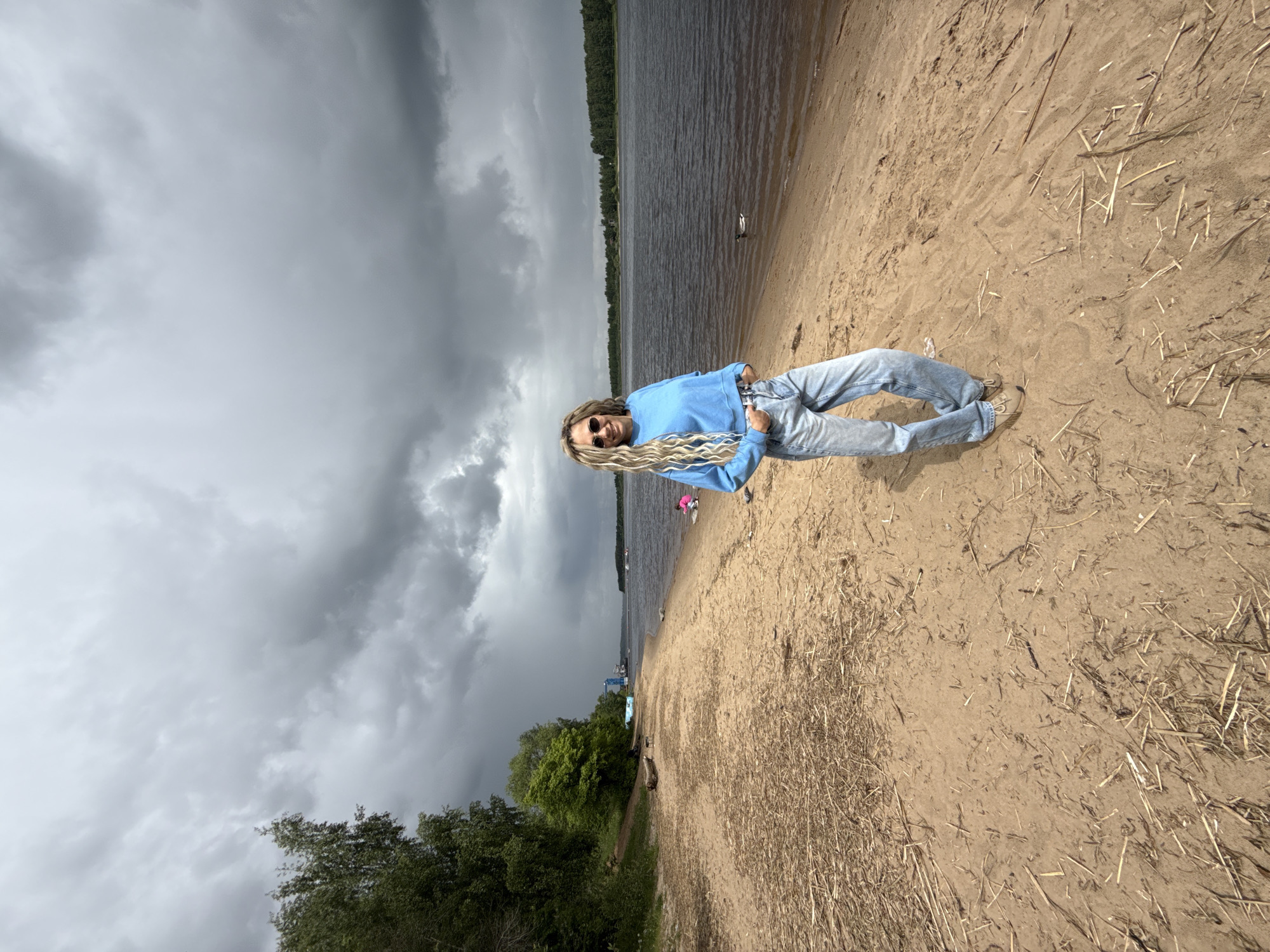 A person wearing sunglasses, a light blue top, and jeans stands on a sandy beach. The location is most likely Rusanovo, Russia, given the provided context. The sky above is overcast with dark, heavy clouds. Behind the person is a body of water, and beyond that, a tree-lined shore. Debris litters the beach.