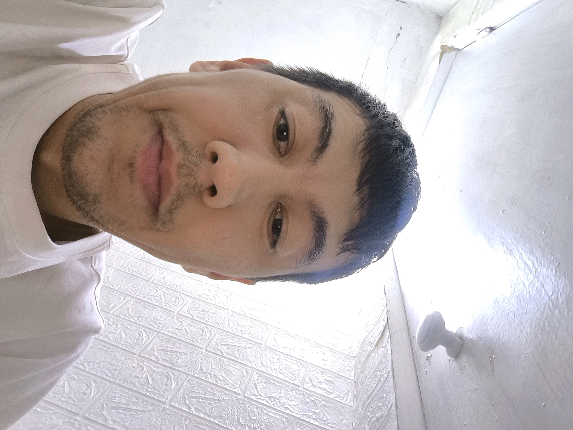 The image is a close-up, low-angle selfie of a person's face, rotated 90 degrees clockwise. The person appears to be male with short, dark hair, light brown eyes, and stubble on his chin and upper lip. He is wearing a white t-shirt. The background consists of textured white walls and a bright, undefined light source. A white doorknob is visible on the right side of the frame. The image's orientation suggests it was taken from a very low perspective, possibly while lying down or looking up from a floor-level position. The location is specified as Kirg Shelk, Kyrgyzstan.
